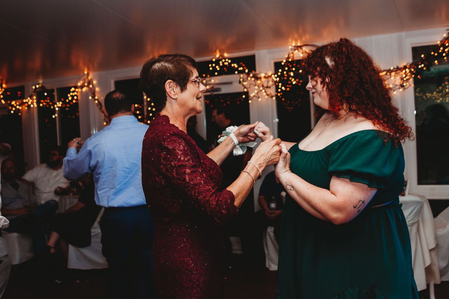 Wedding guests dancing together at a reception with string lights and red lighting creating a festive atmosphere.