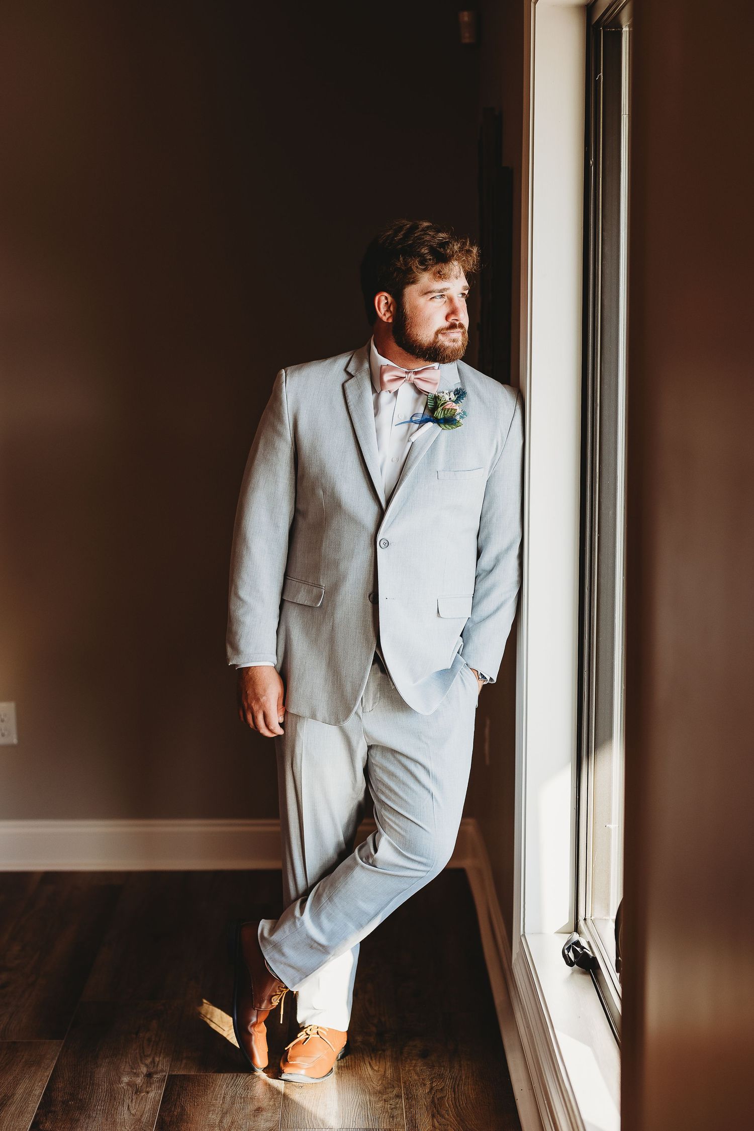 Groom poses by window in light gray suit for stylish wedding day portraits.