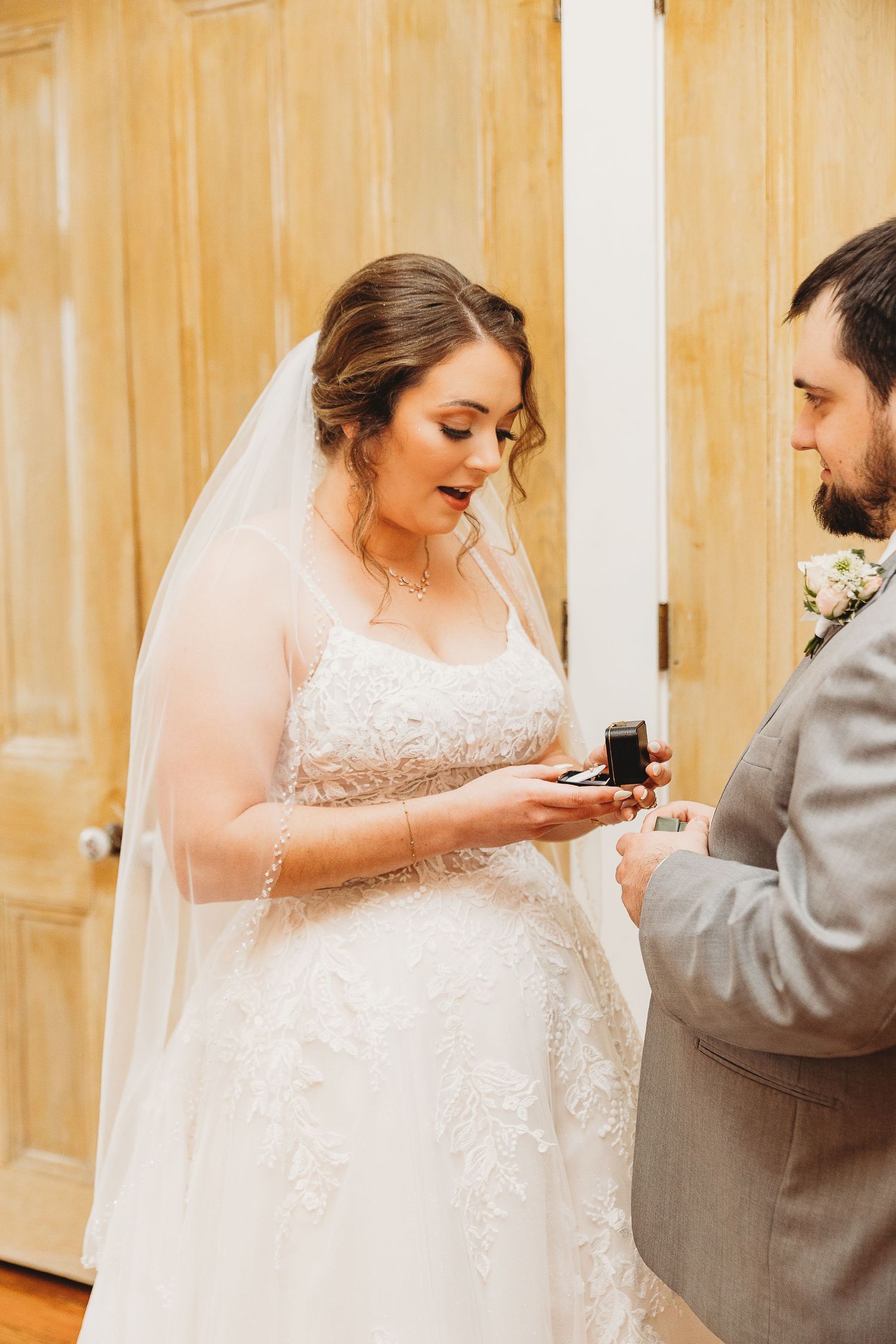 A bride in a lace wedding dress and veil examines a ring box during an intimate ceremony moment.
