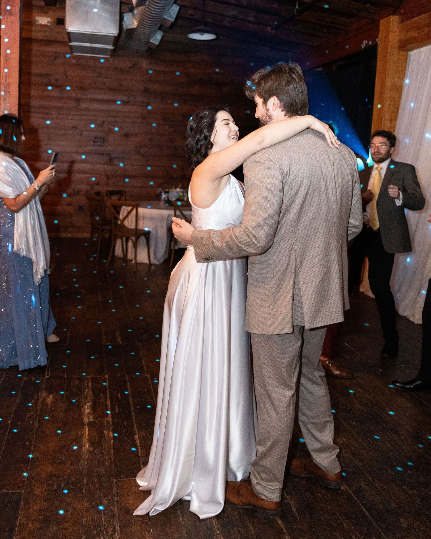 A couple dancing at a wedding reception in a rustic wooden venue with blue confetti falling around them.