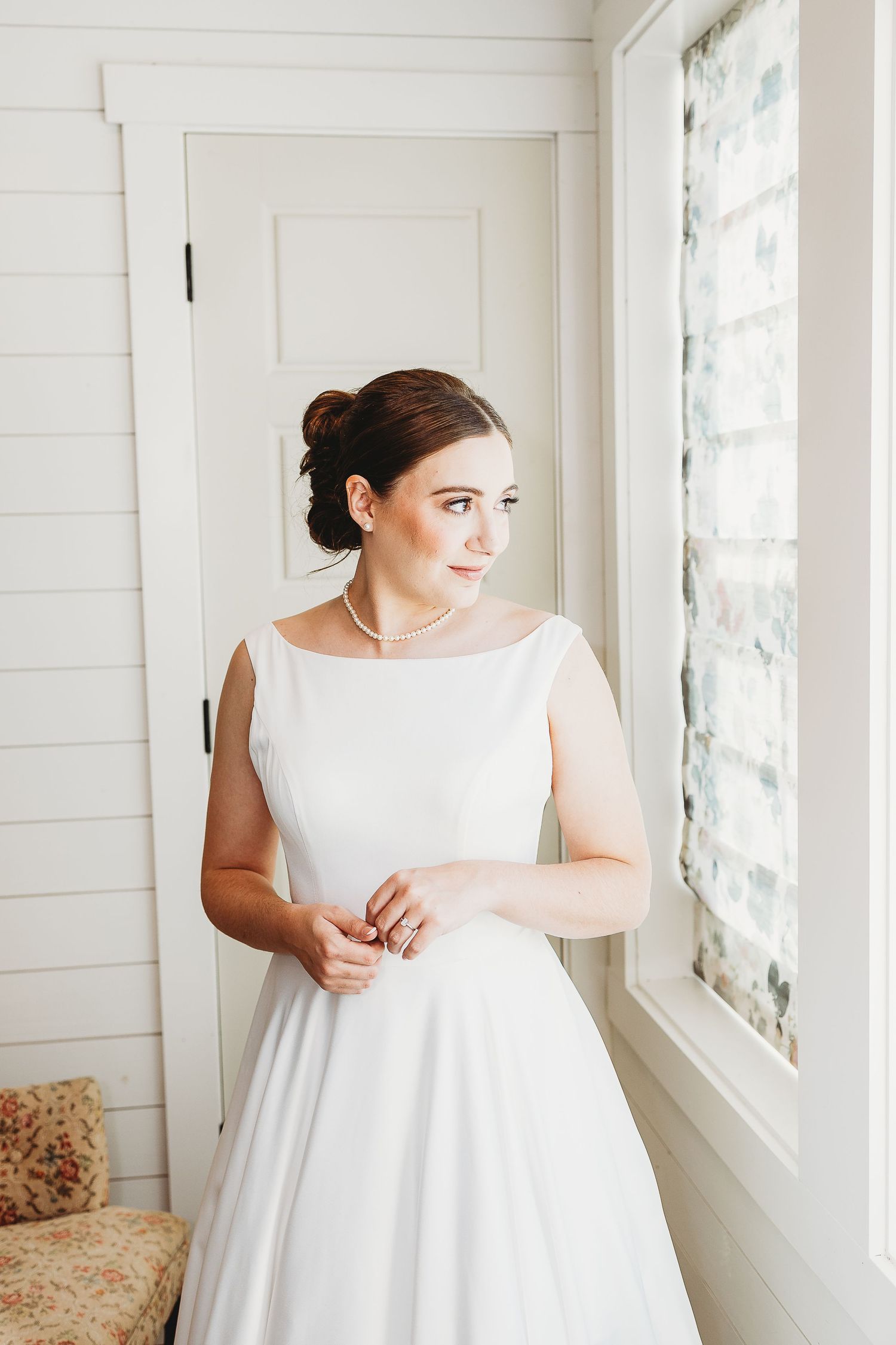 A bride in a classic white wedding dress stands by sunlit windows in an elegant interior.