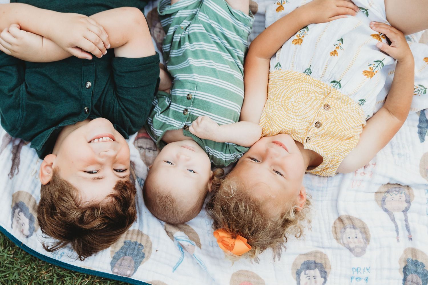 Three children lying on a patterned blanket outdoors.