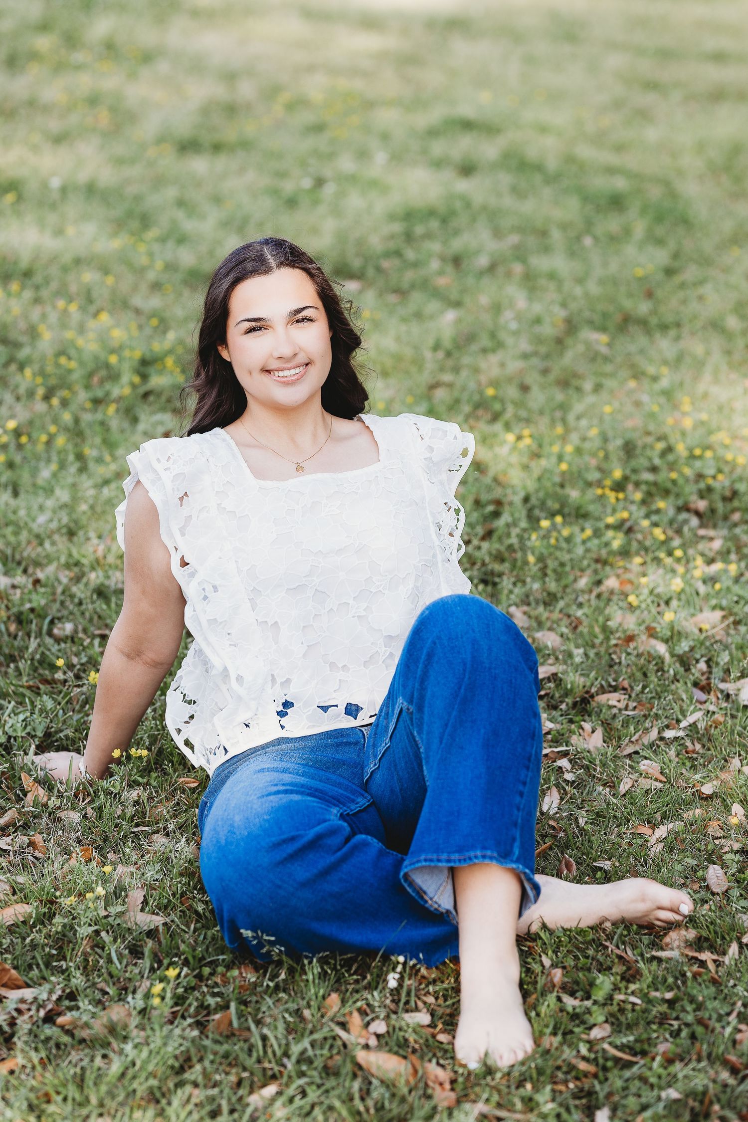 Casual outdoor portrait showing white lace top and blue jeans while sitting in a grassy field.