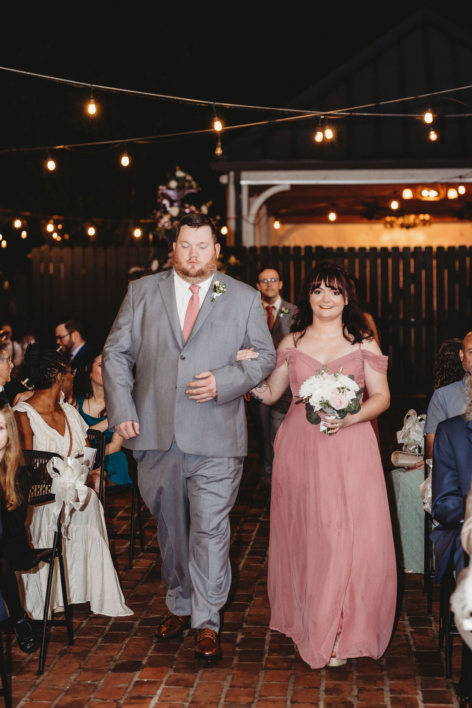 Wedding party members walk down a brick aisle under romantic string lights at dusk.