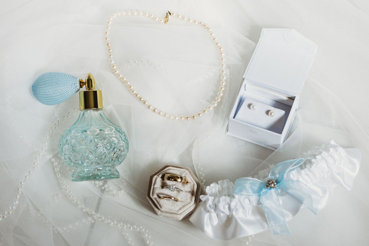Wedding accessories including a blue perfume bottle, pearl necklace, and garter on white surface.