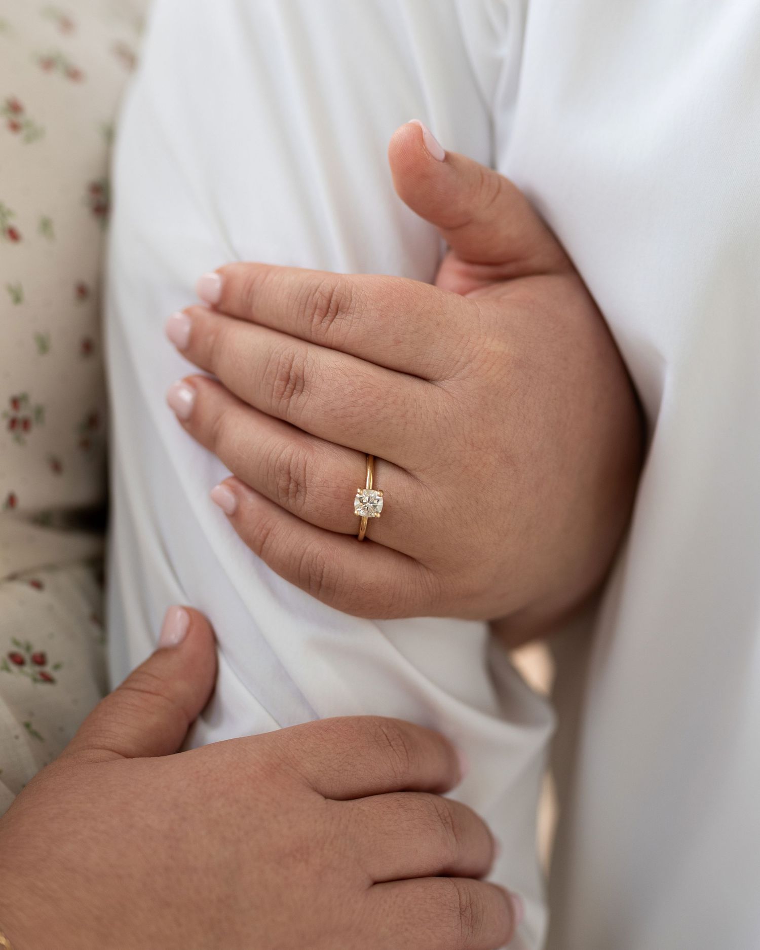Close-up view of hands wearing an engagement ring against a white fabric background.