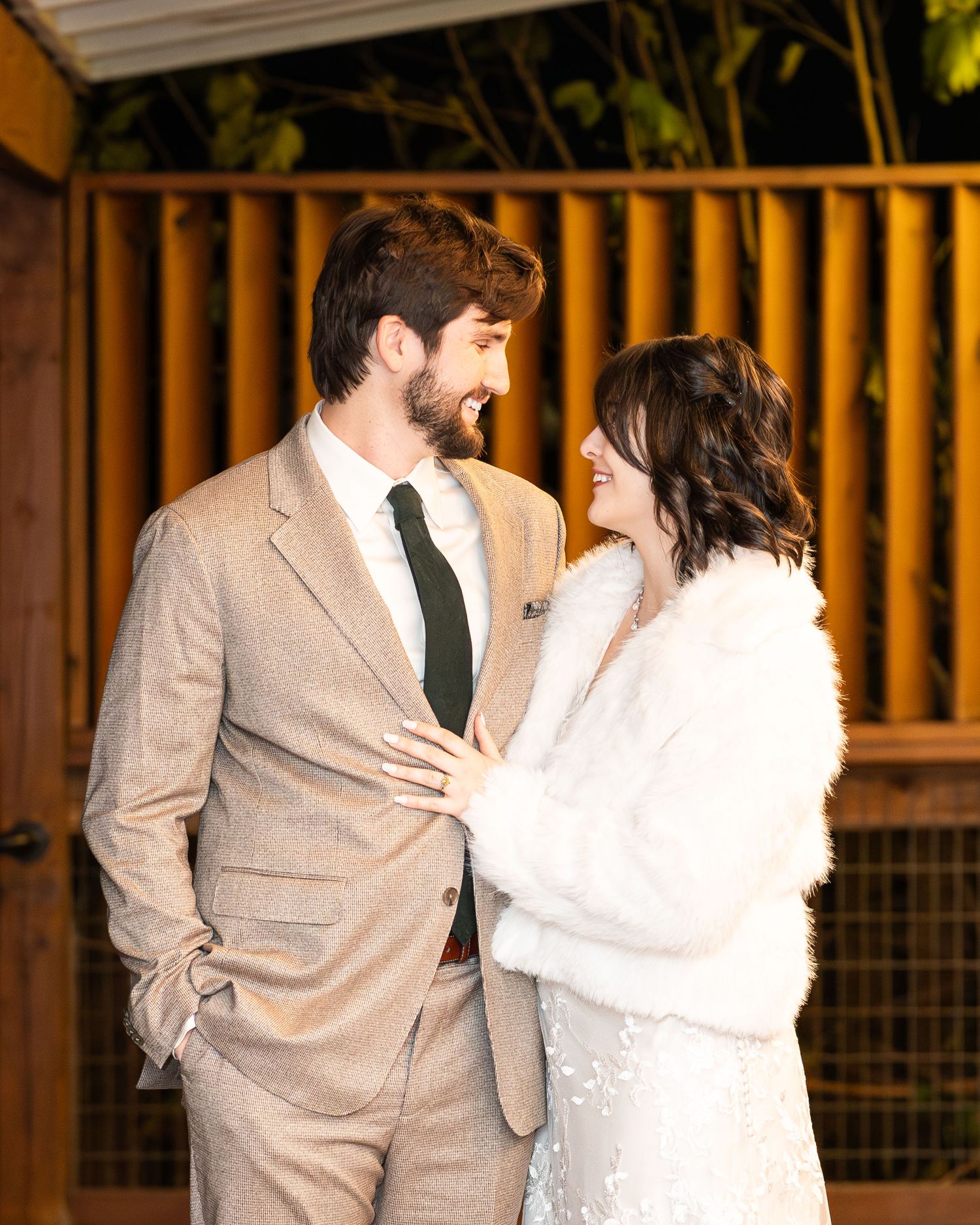 A couple in wedding attire share an intimate moment against a wooden fence backdrop at sunset.