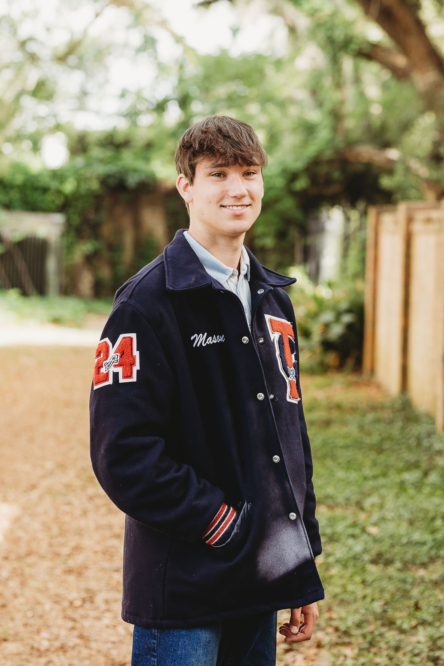 Student in navy varsity jacket with number 24 smiles on tree-lined path.