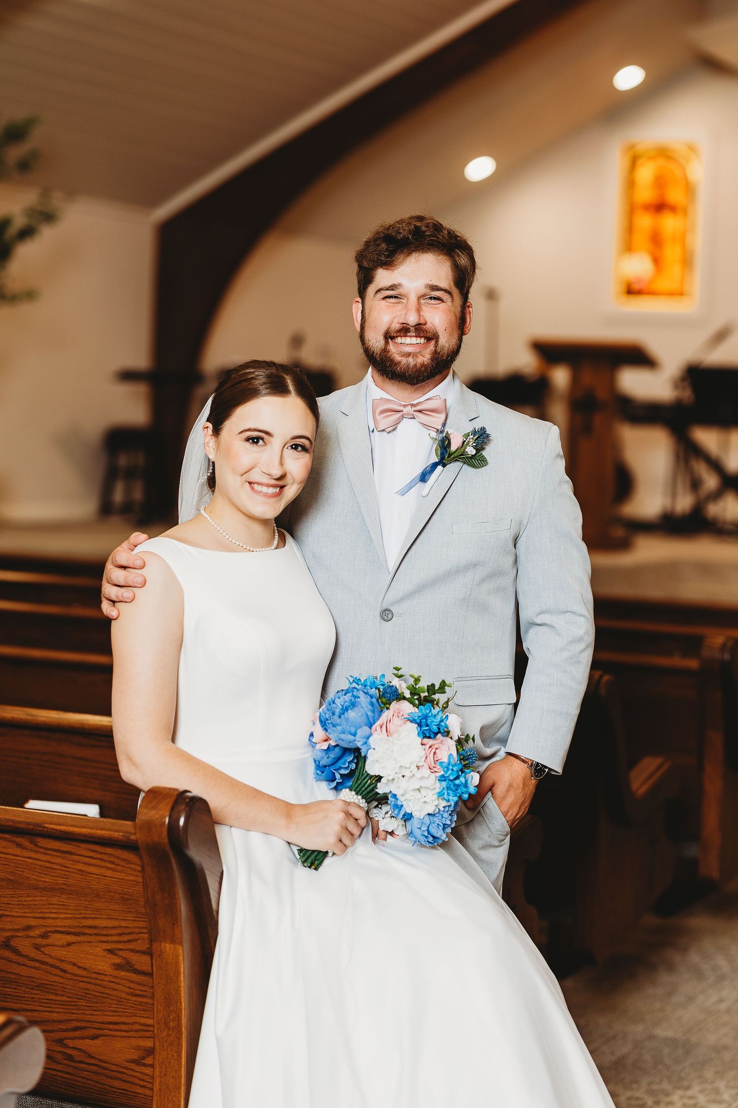 A couple in wedding attire poses together holding a blue and white floral bouquet in an indoor setting.
