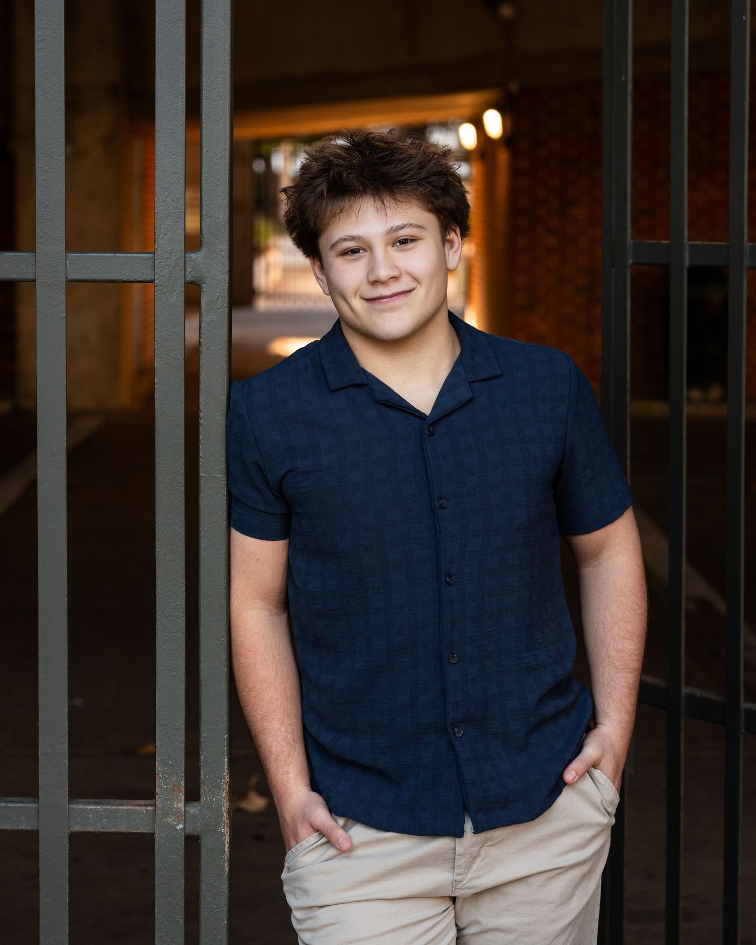 Young person wearing a navy blue polo shirt and khaki pants poses outdoors against a brick wall background.