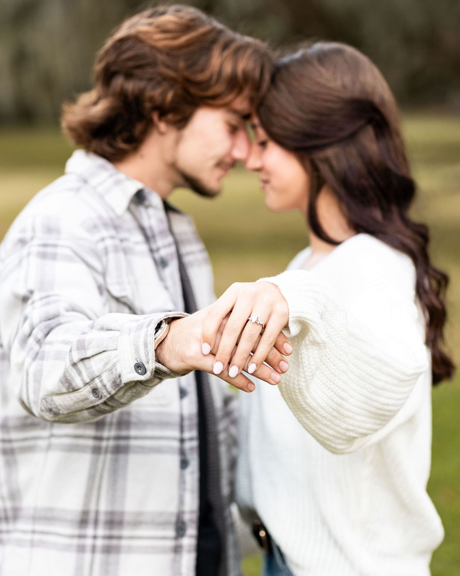 A couple in casual winter attire shares an intimate moment while showing off an engagement ring outdoors.