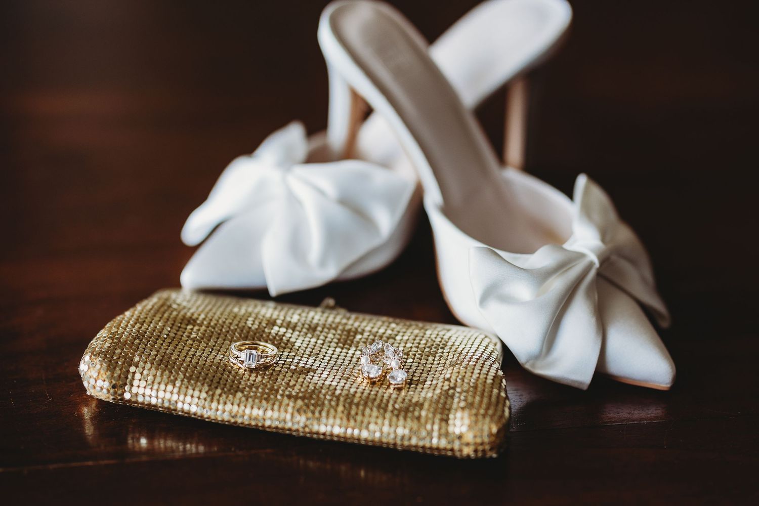 Gold sequined clutch purse and white bow accessories for a wedding on dark wooden surface.