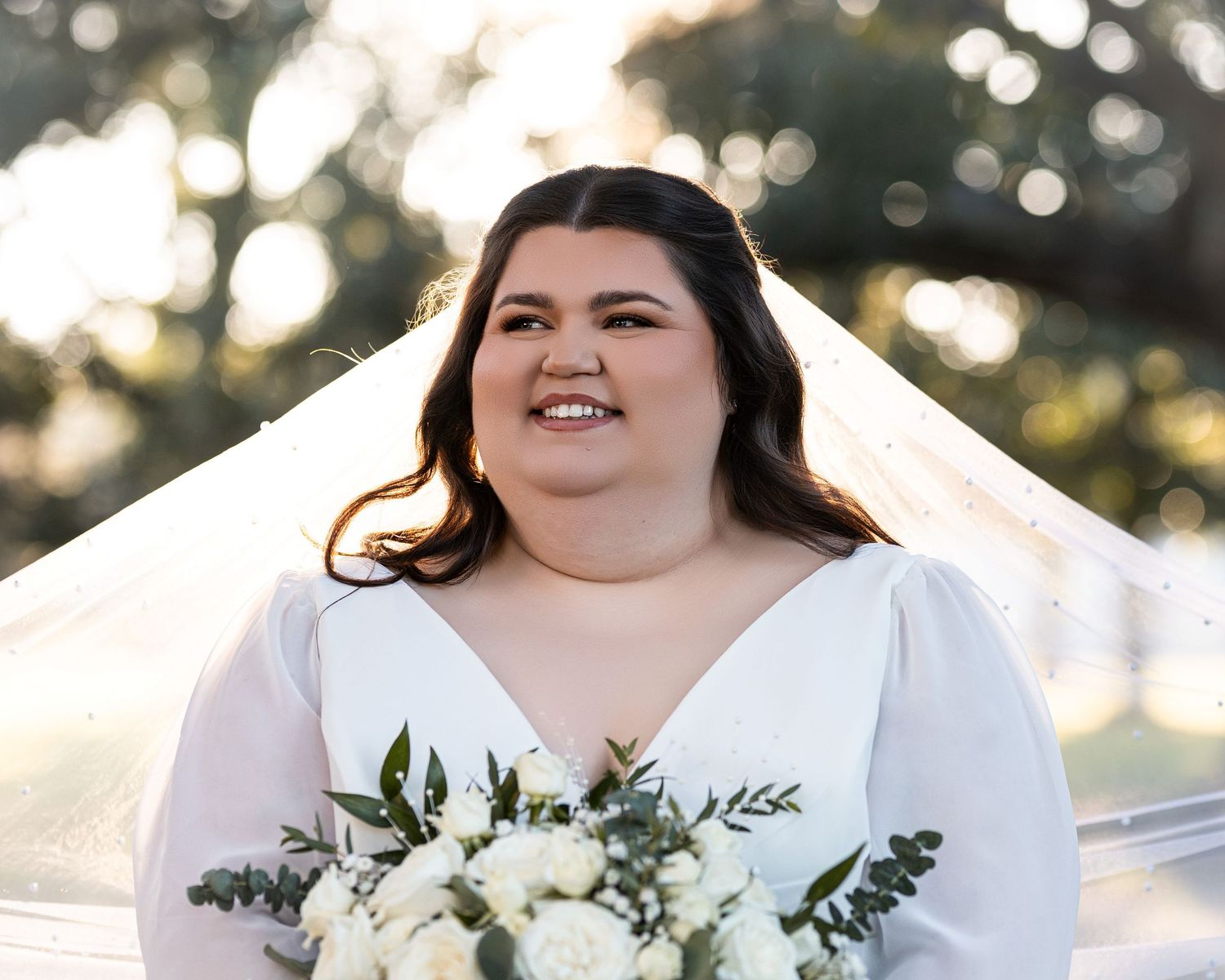A bride in a white wedding dress holds a lush bouquet of white flowers and greenery during an outdoor ceremony.
