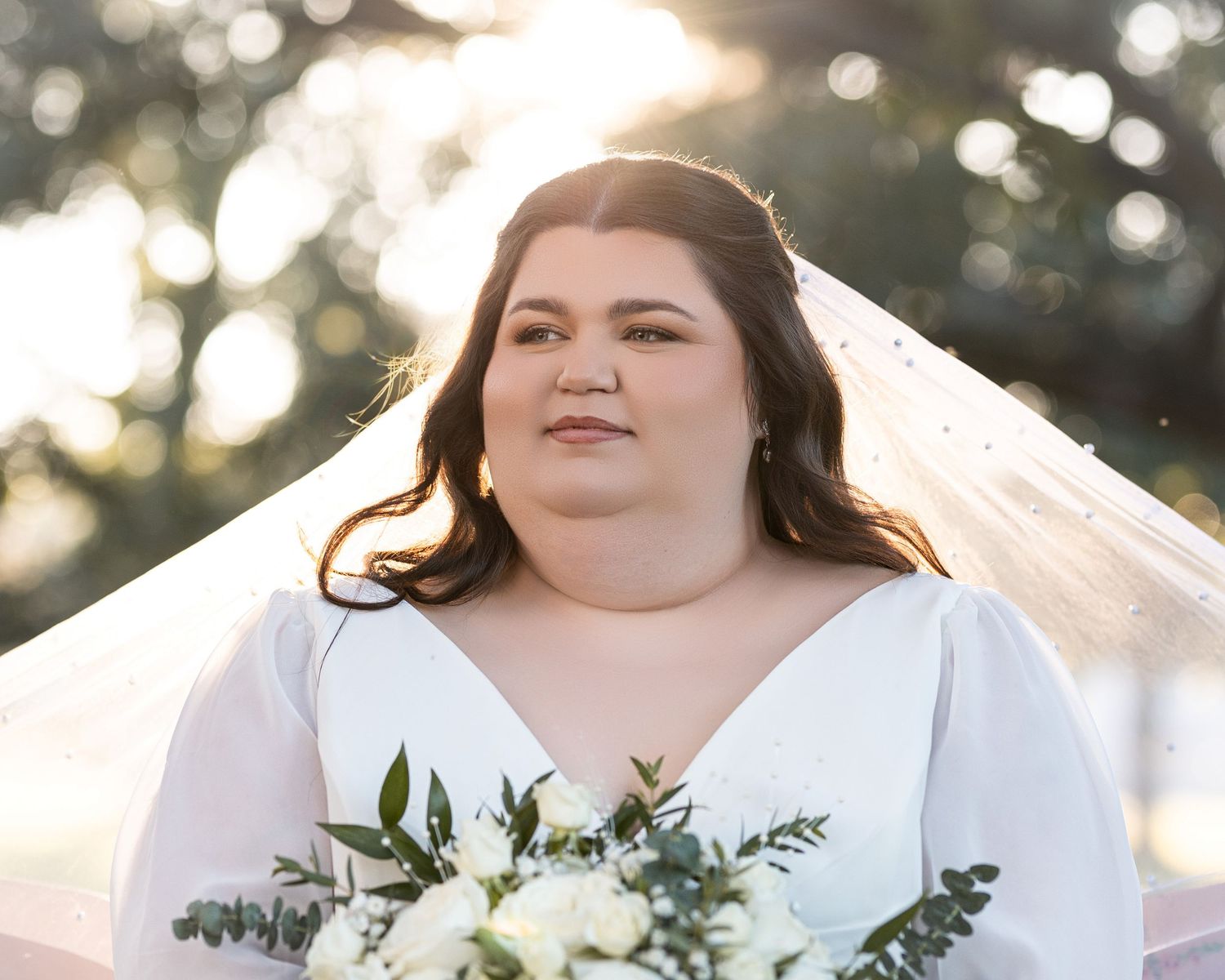 A bride in a white wedding dress holds a delicate bouquet of white and green flowers during a sunlit outdoor photo session.