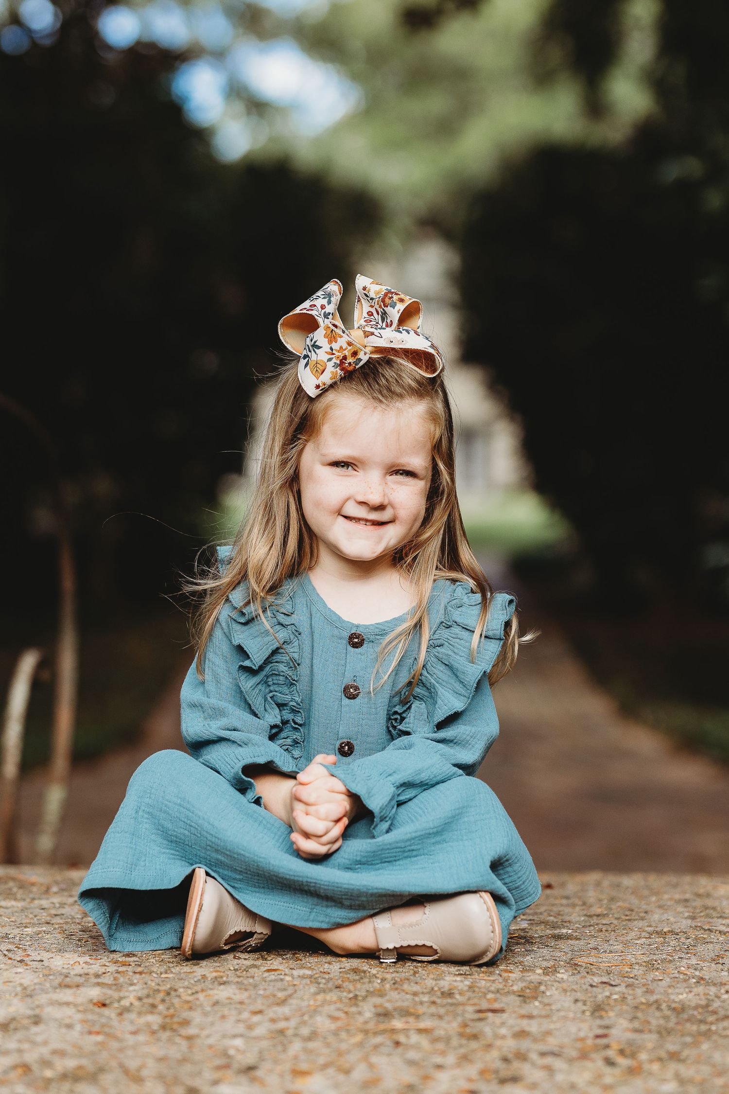 Child in teal dress sitting cross-legged on ground wearing floral hair bow.