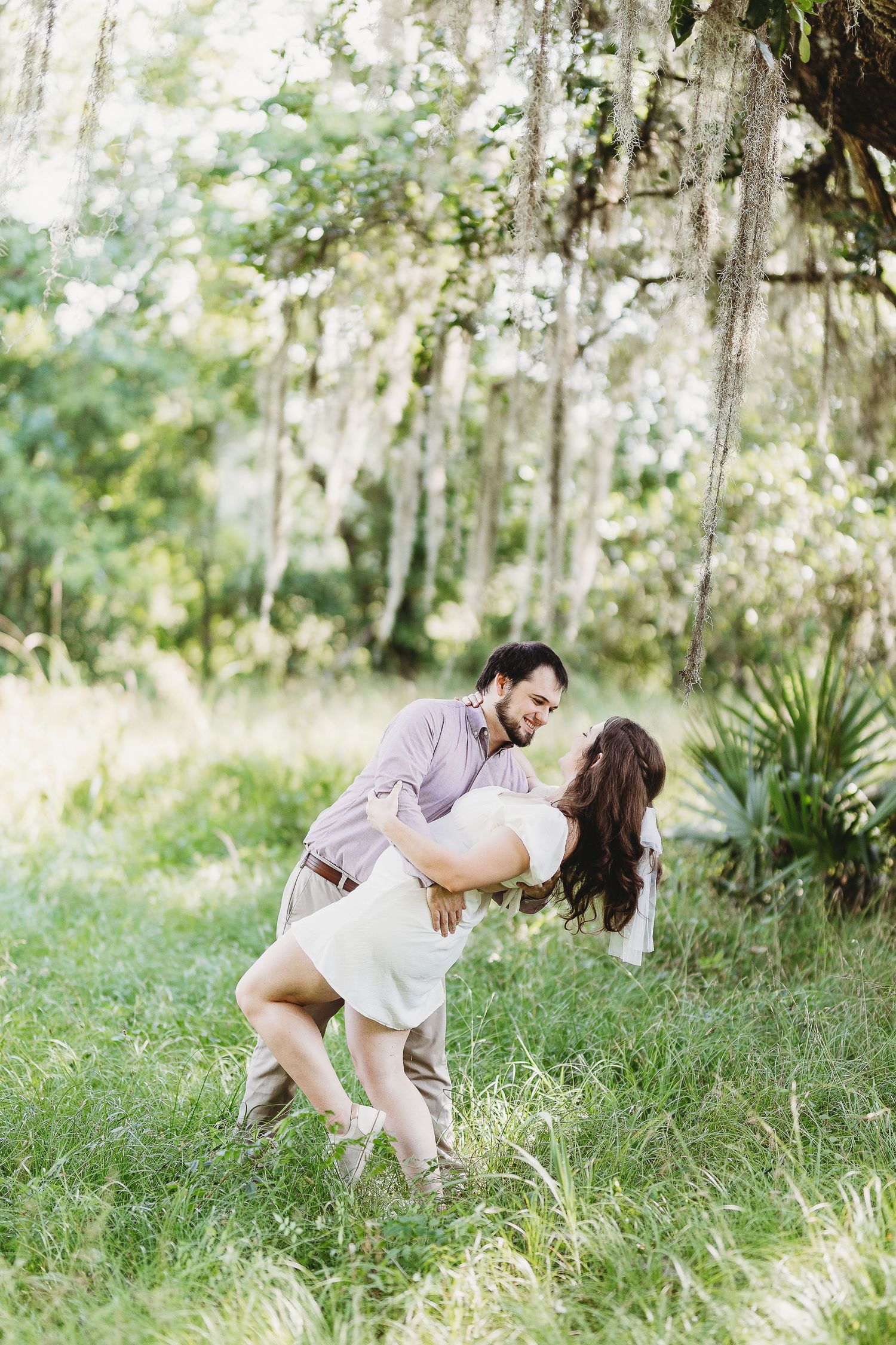 A couple dances and spins playfully in a sunlit meadow with Spanish moss in the background.