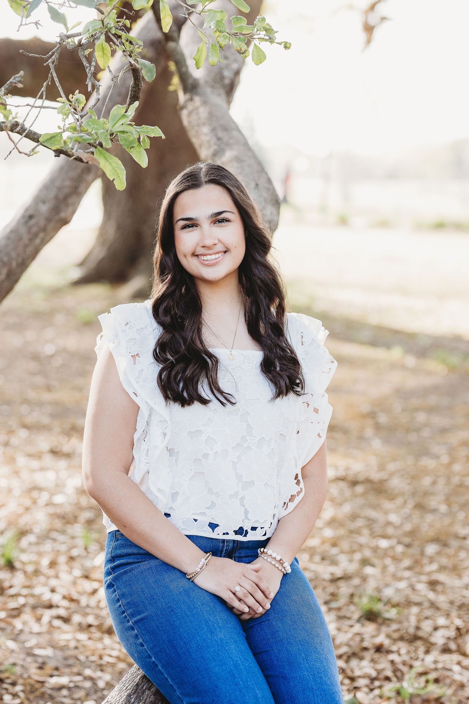 Person in white lace top sits outdoors under tree branches.