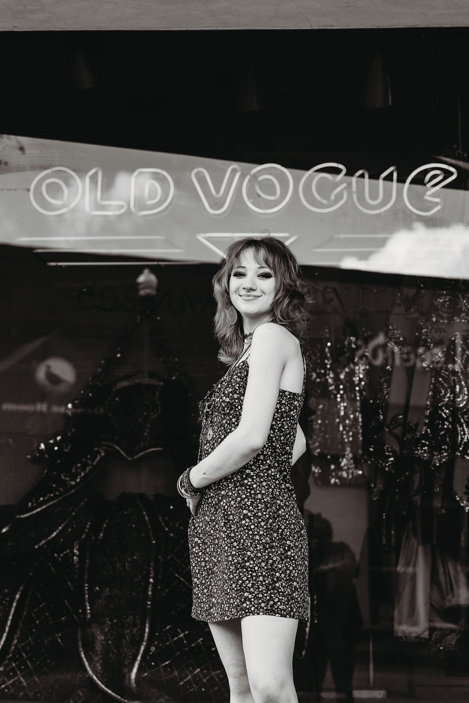 A black and white portrait in front of an Old Vogue storefront sign wearing a patterned dress.