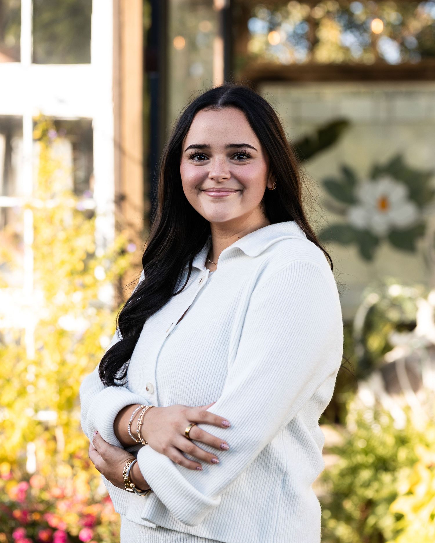 Professional portrait of someone in a white sweater smiling warmly outdoors with autumn foliage in the background.