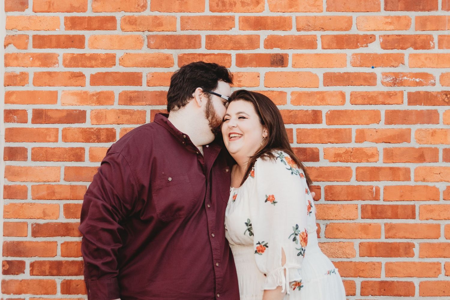 Romantic couple poses against rustic brick wall during outdoor photo session.