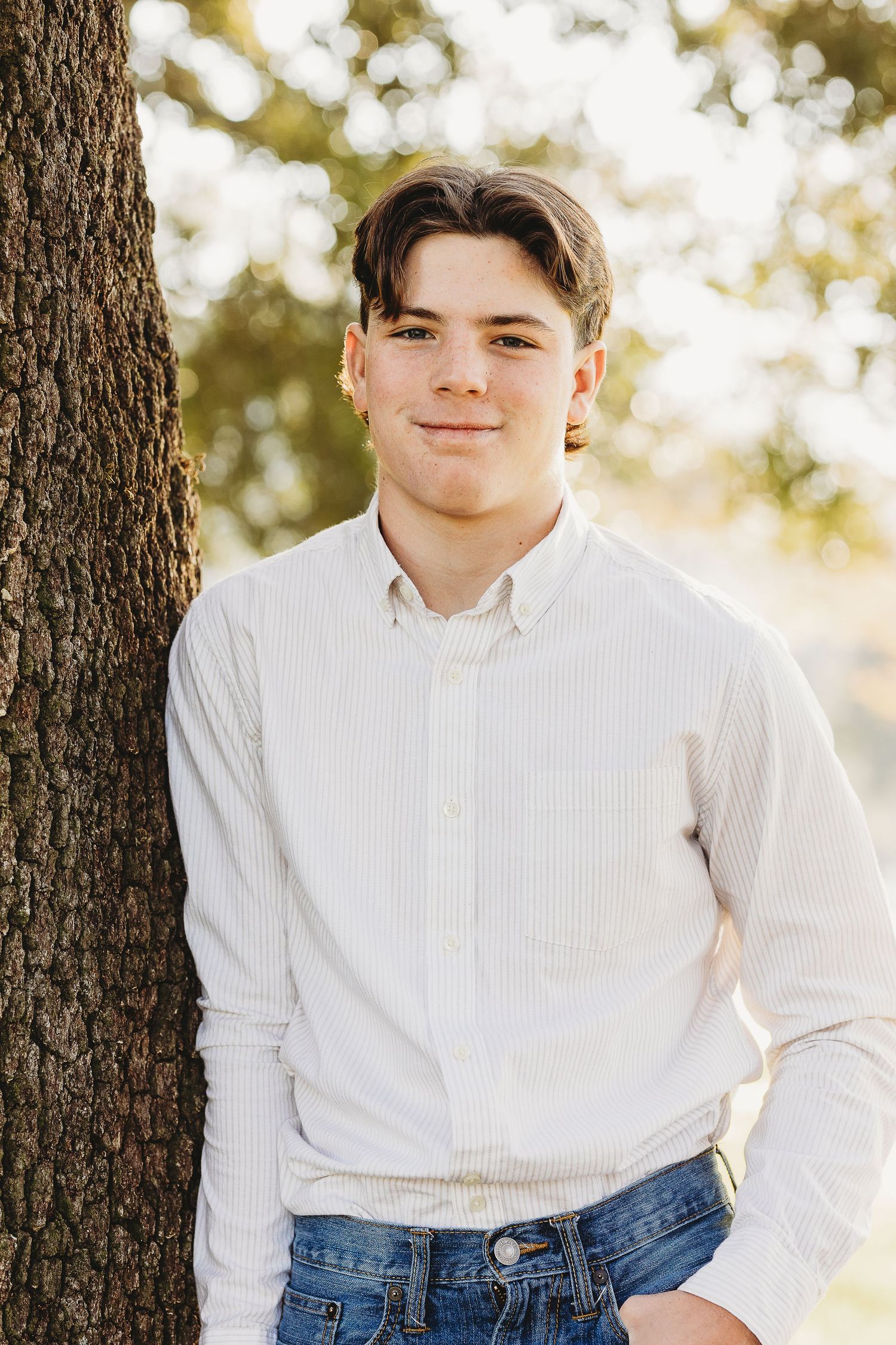 Portrait series against tree trunks wearing white button-down shirt and jeans.