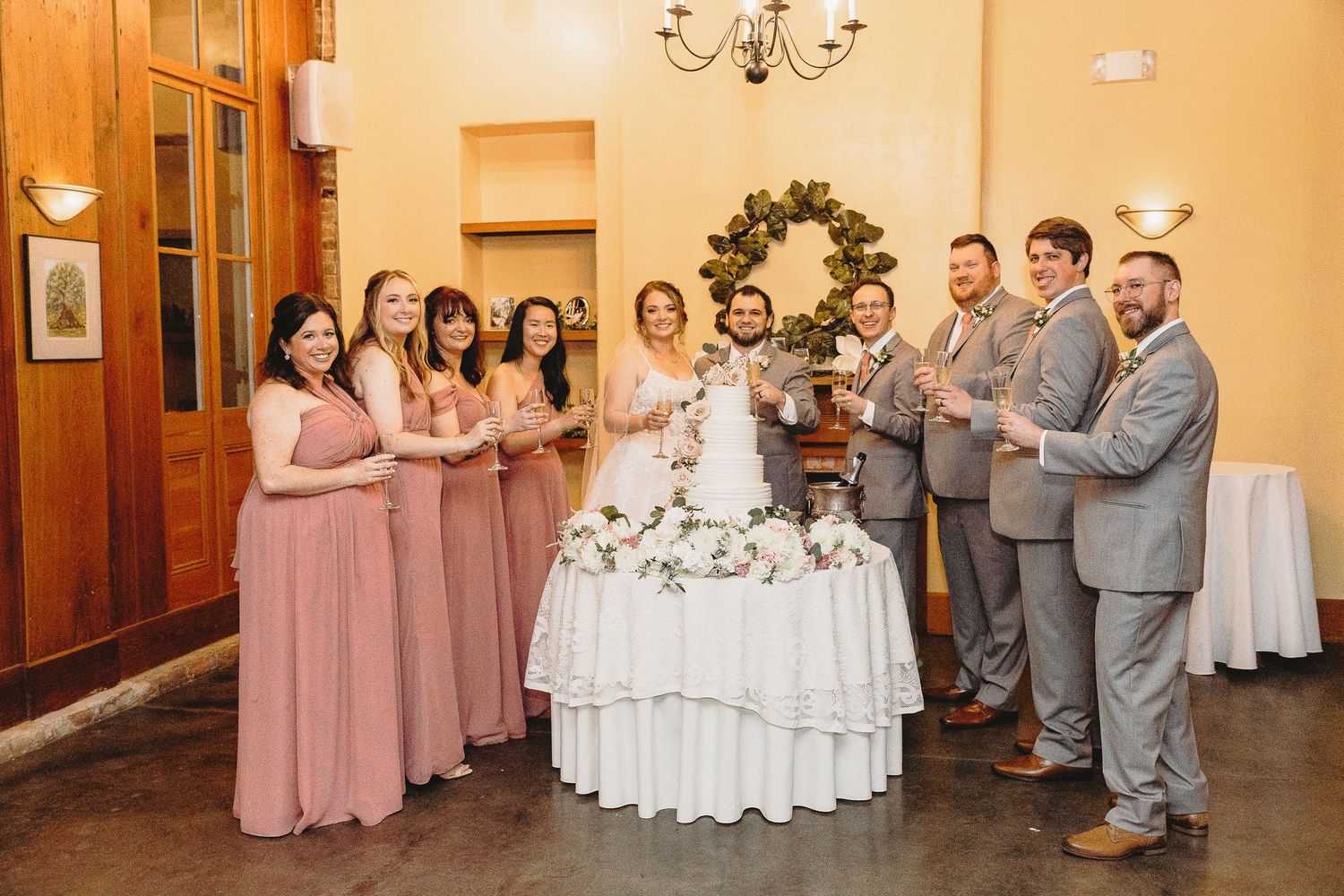 Wedding party poses in a reception hall decorated with chandeliers and pink bridesmaid dresses.