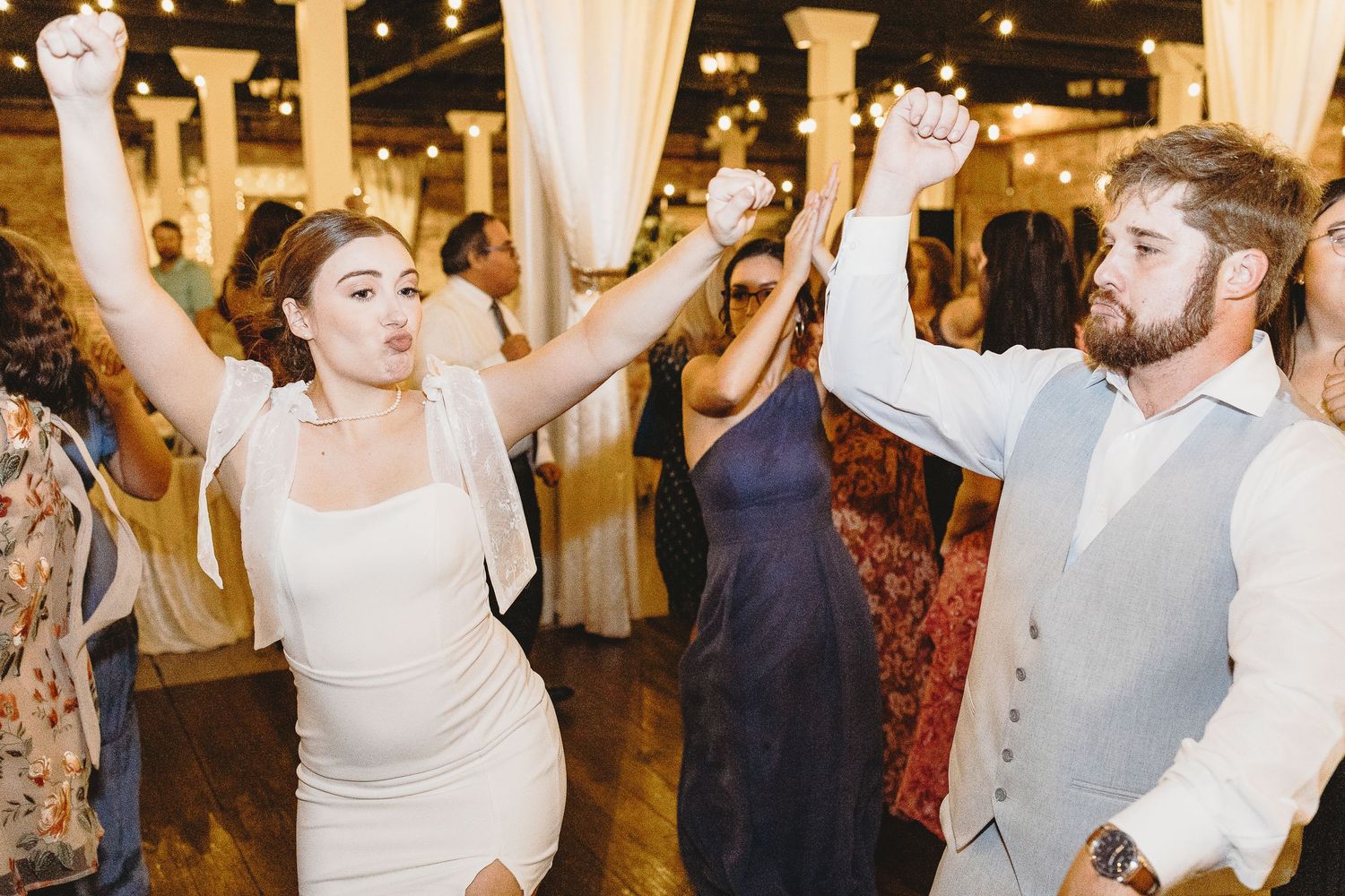 Wedding guests dancing and celebrating at an indoor reception with white drapes and lighting.
