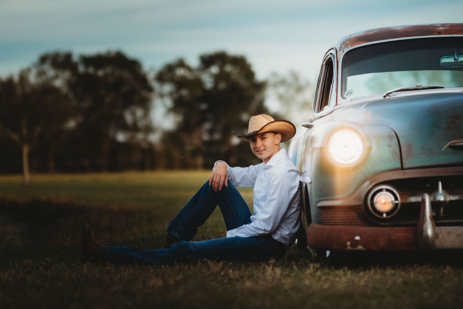 A vintage-style portrait of someone sitting beside a classic car at sunset.