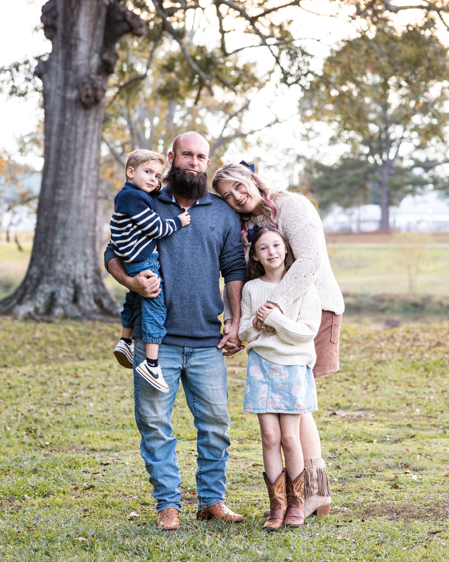 A casual family portrait taken outdoors with a rustic tree backdrop on a sunny day.