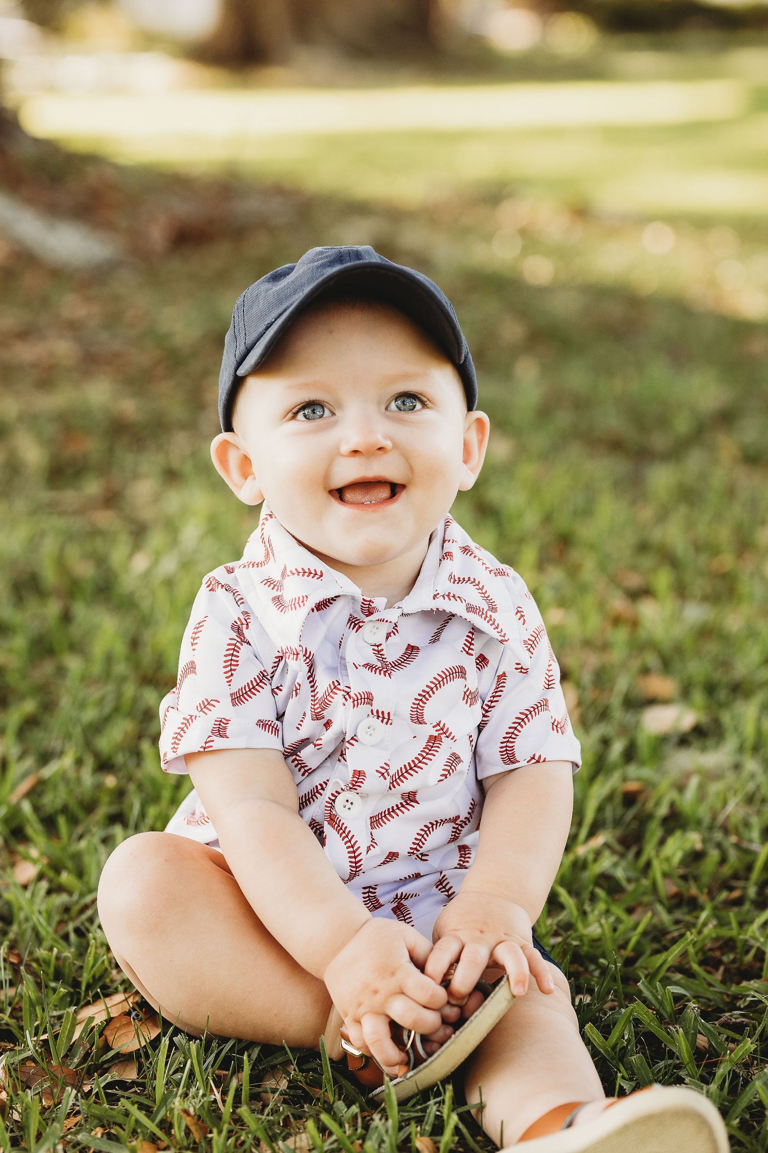 A baby wearing a baseball cap and patterned shirt sits smiling on grass during a sunny day.