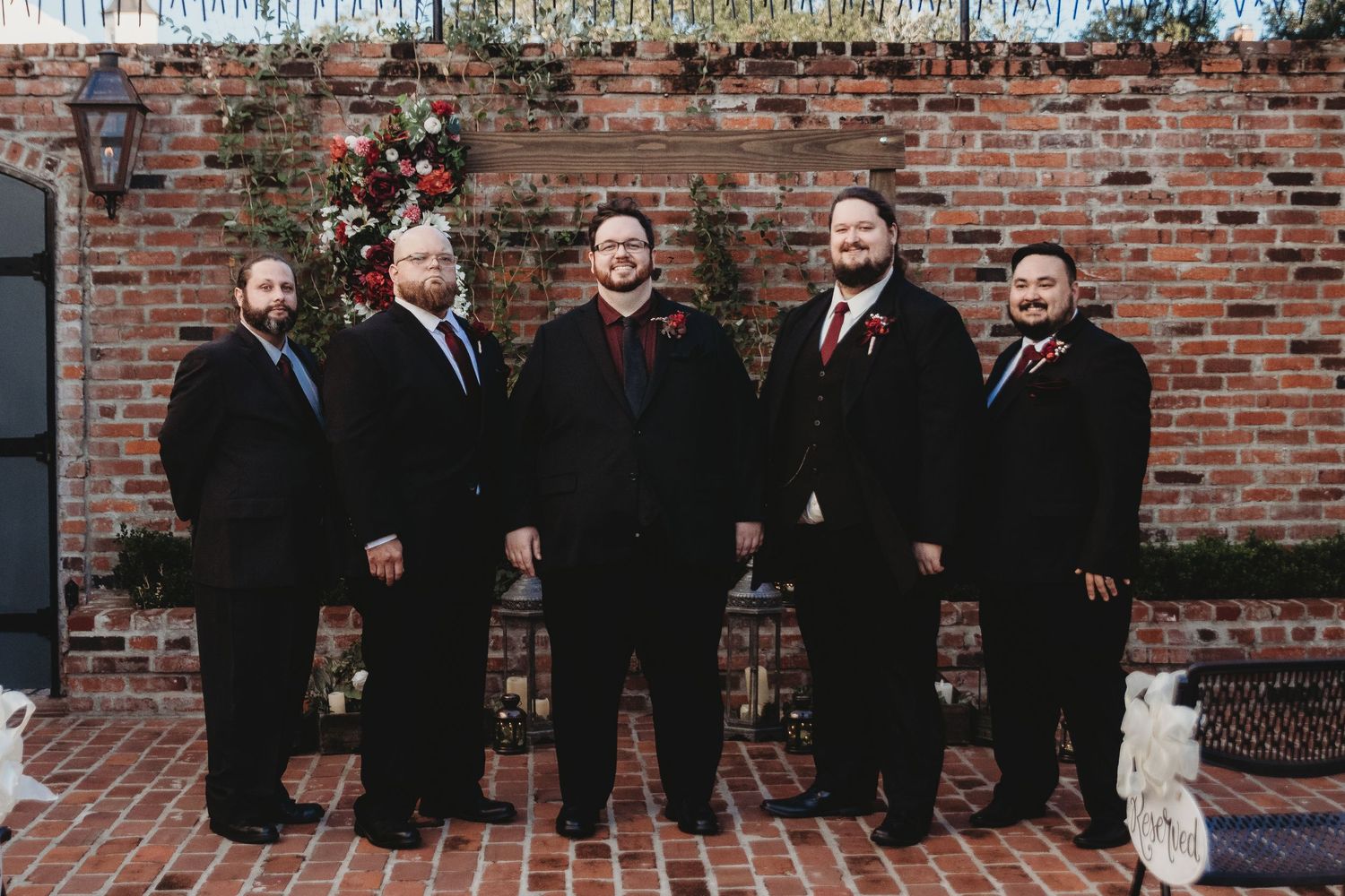 Groomsmen in black suits pose together against a rustic brick wall backdrop.