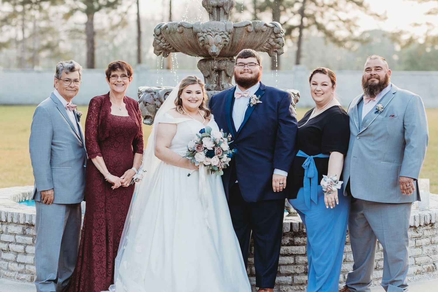 Wedding party poses for formal photos in front of a stone fountain on a sunny afternoon.
