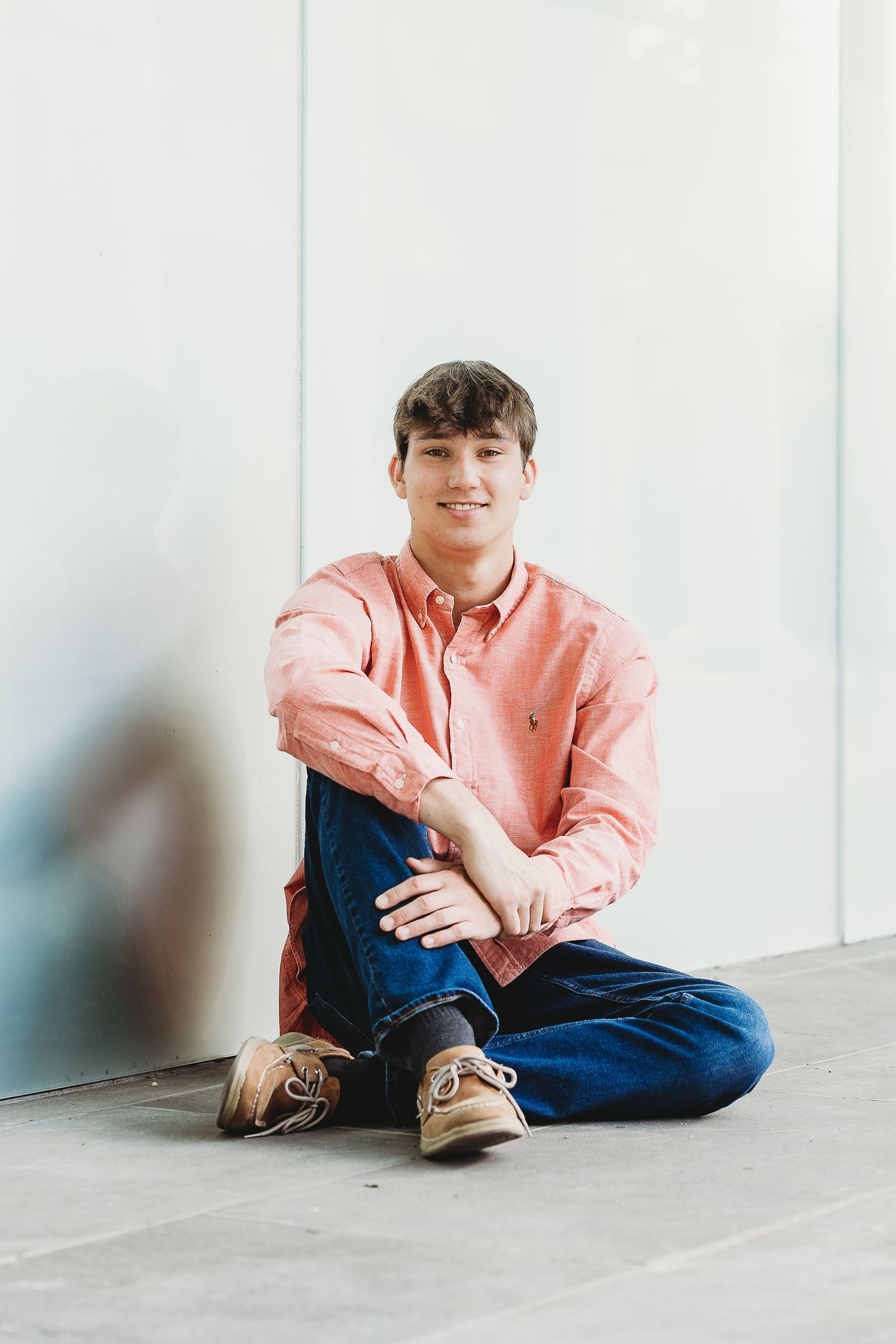 Casual portrait of someone in salmon colored button-down shirt sitting against white wall.