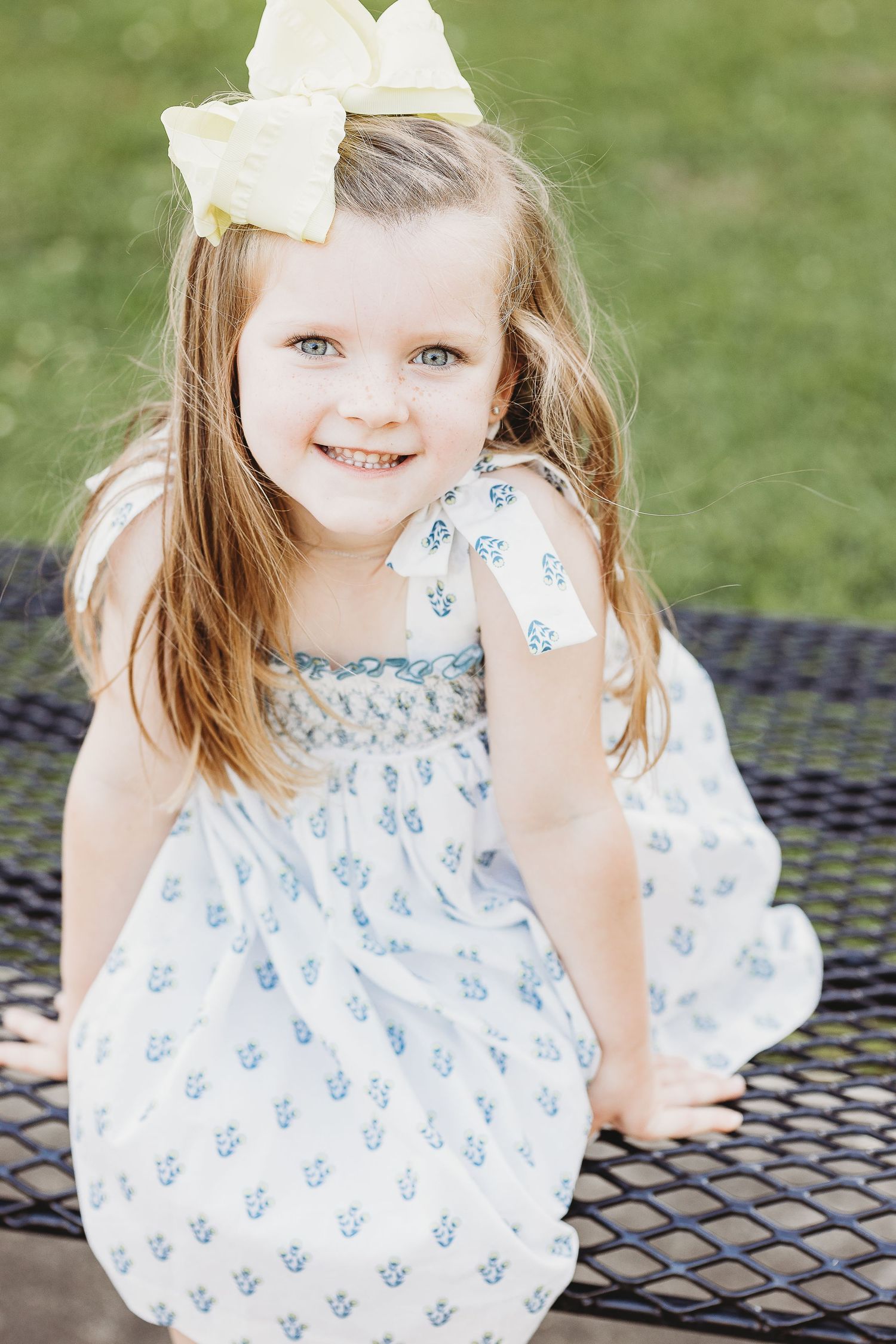 A child in a white dress with blue patterns and bow poses outdoors on metal furniture.