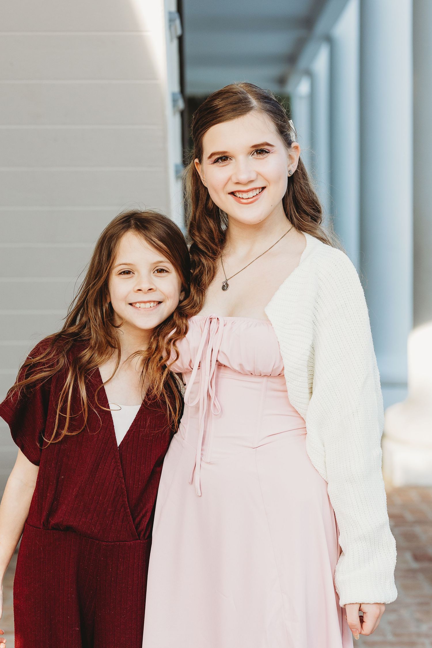 Two young people in coordinating burgundy and pink dresses pose together against a white column background.