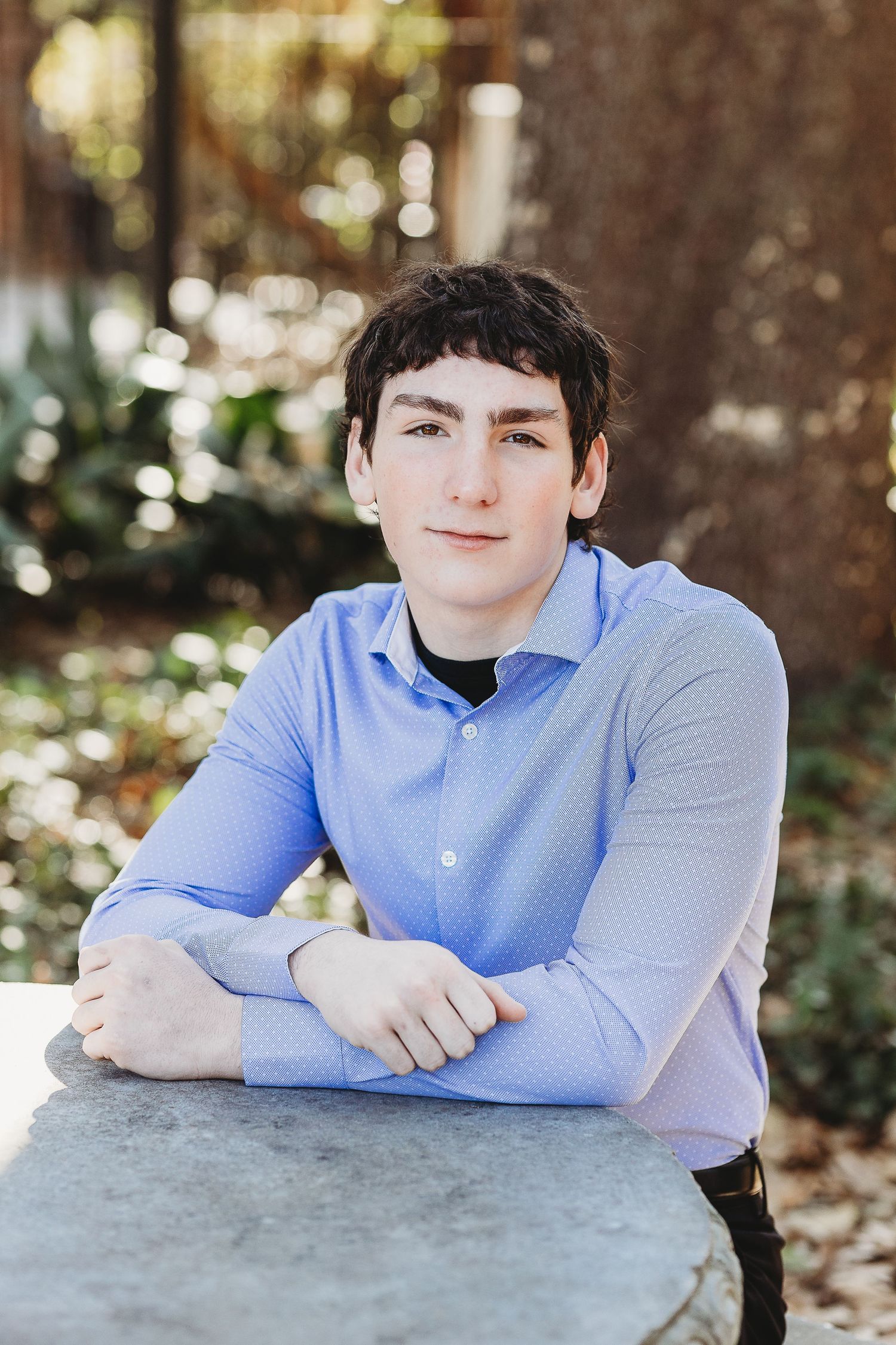 A professional portrait of someone in a light blue button-down shirt leaning on a stone surface.