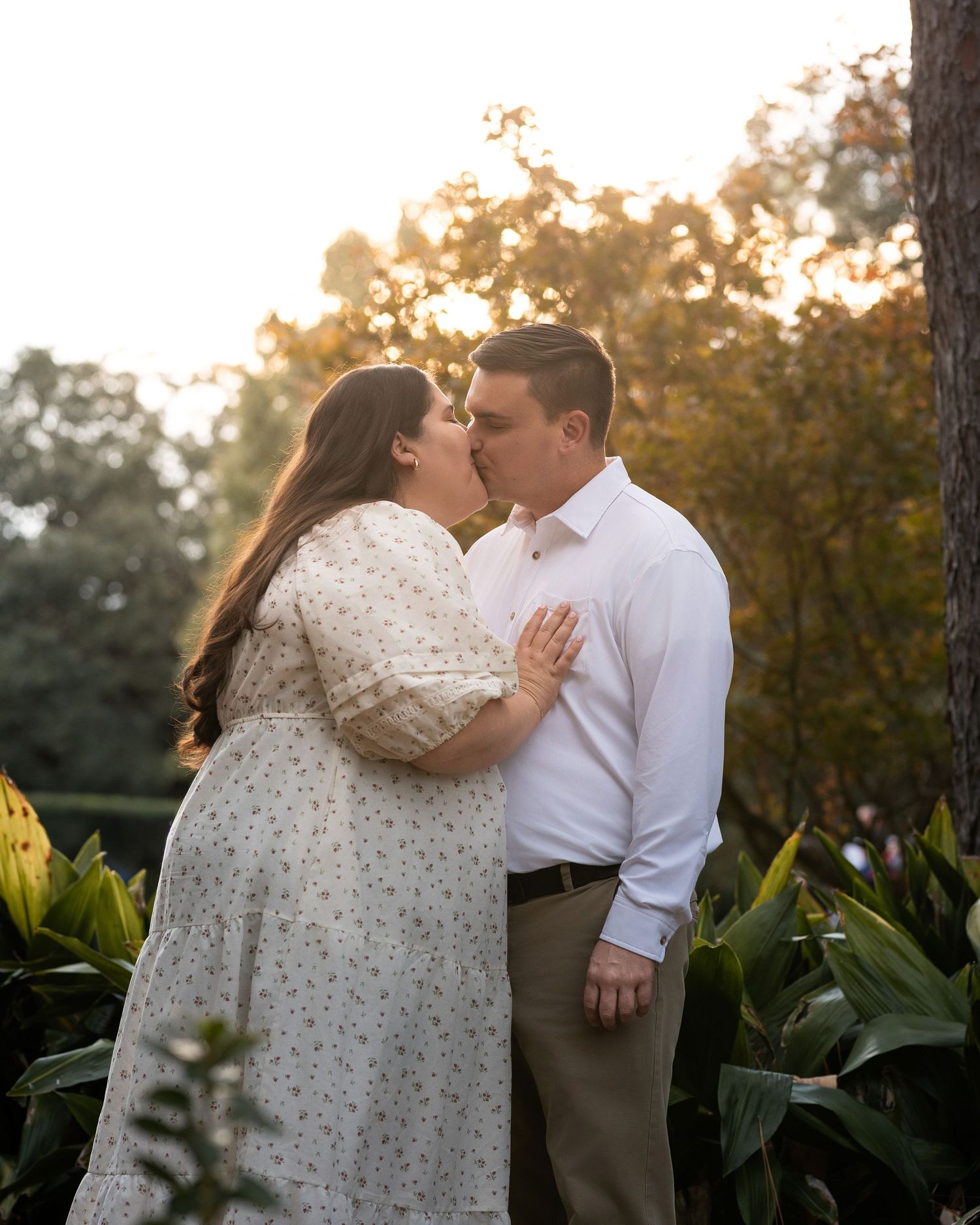 A romantic couple shares a tender kiss in a garden setting during golden hour with lush foliage in the background.
