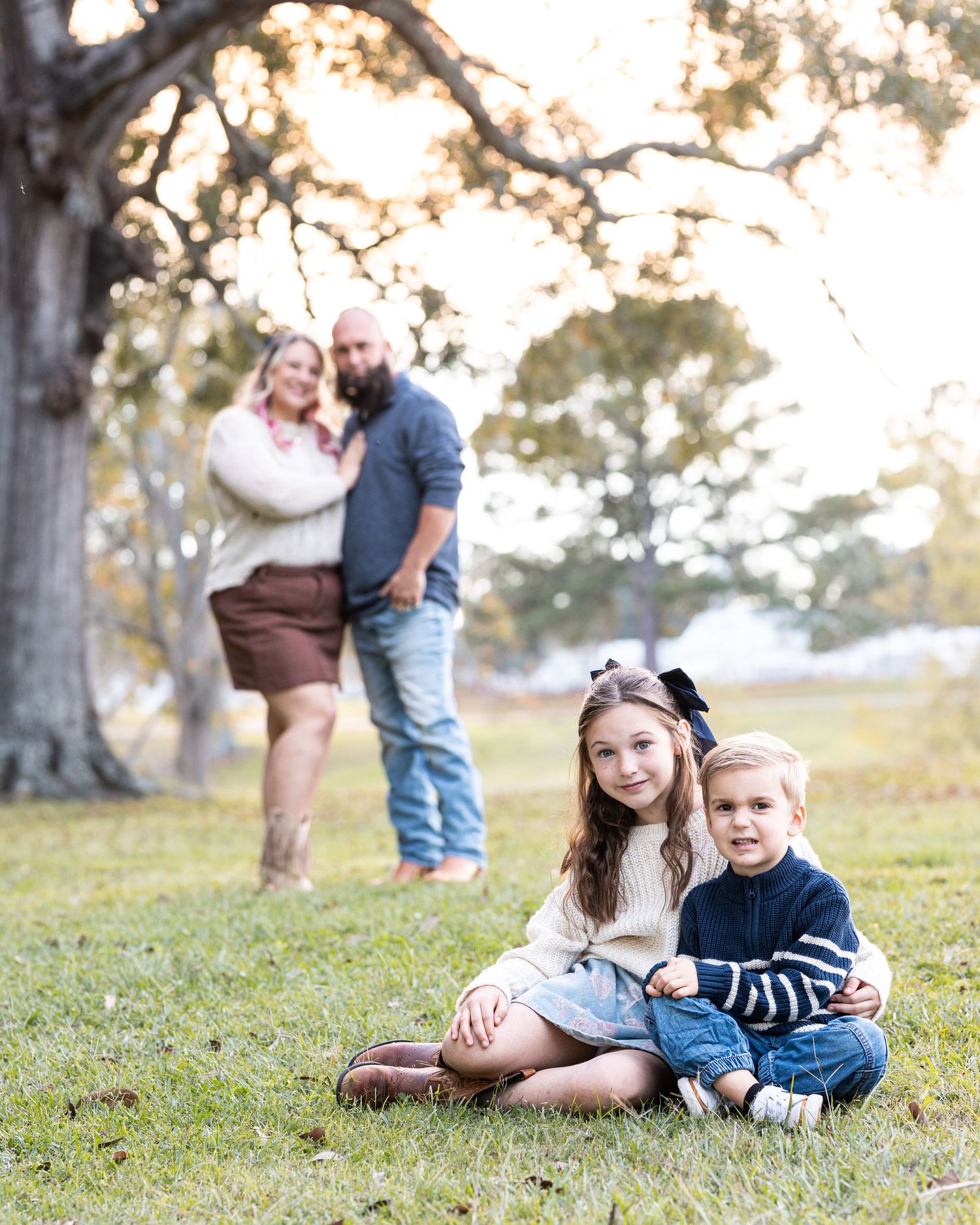 A couple stands in the background while two children sit together on grass under a tree during an autumn photoshoot.