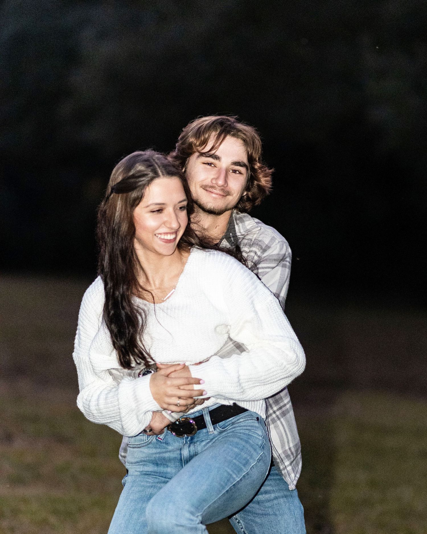 A couple embraces outdoors in a candid moment, with the woman wearing a white sweater and jeans against a dark background.