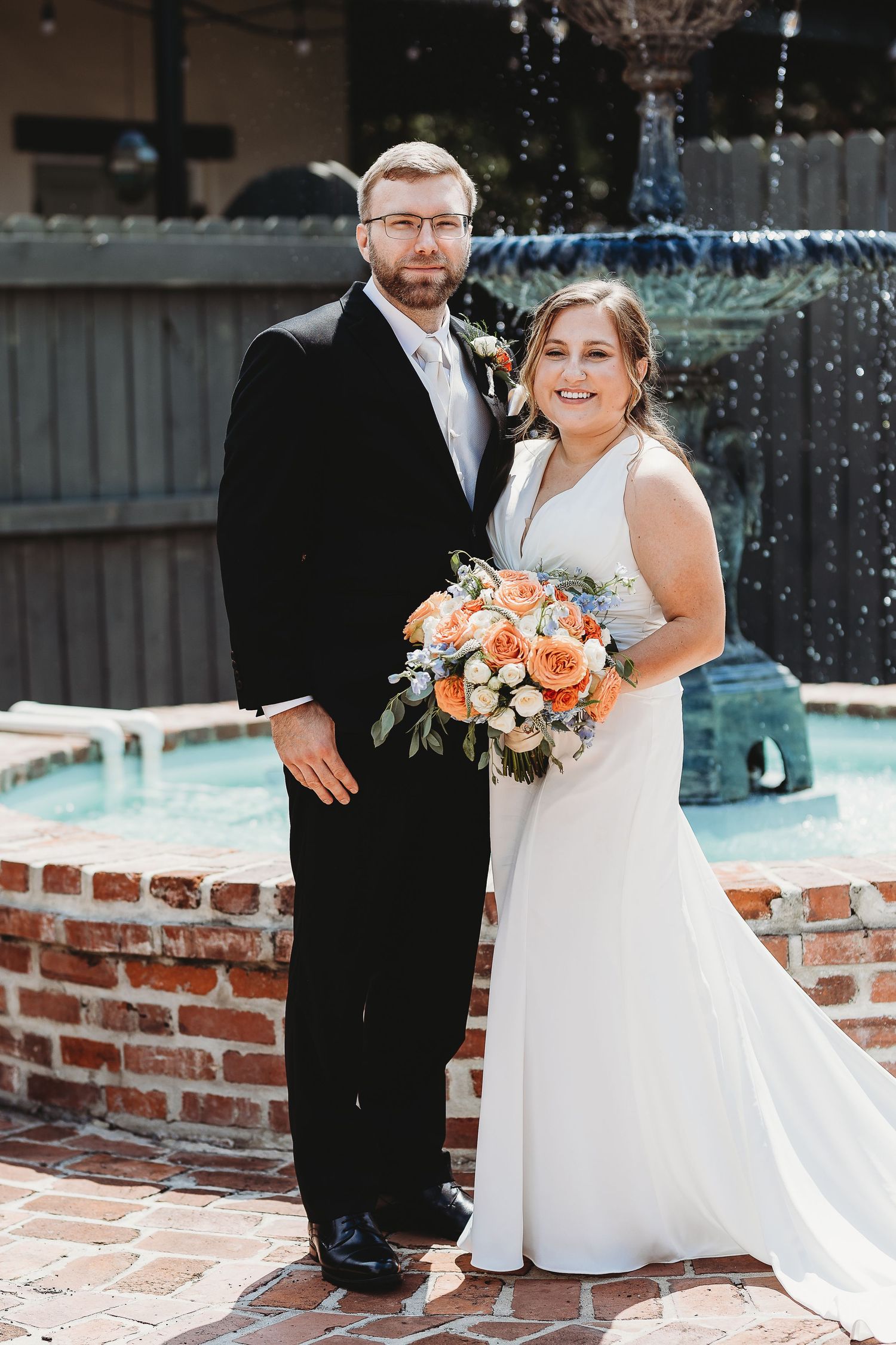 Wedding couple posing by brick wall with peach and white floral bouquet.