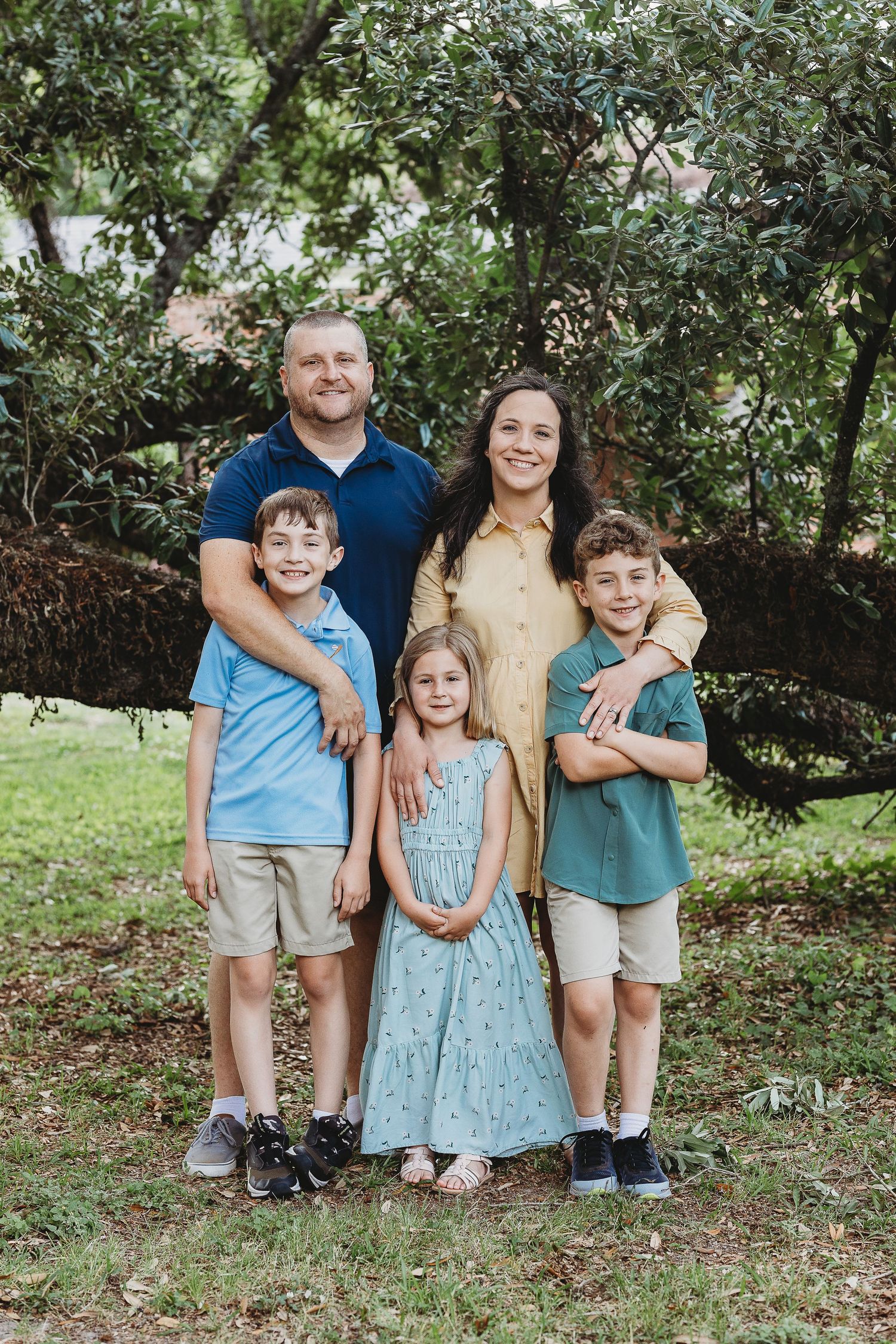 A family poses together under a large tree in a garden setting during a summer photoshoot.