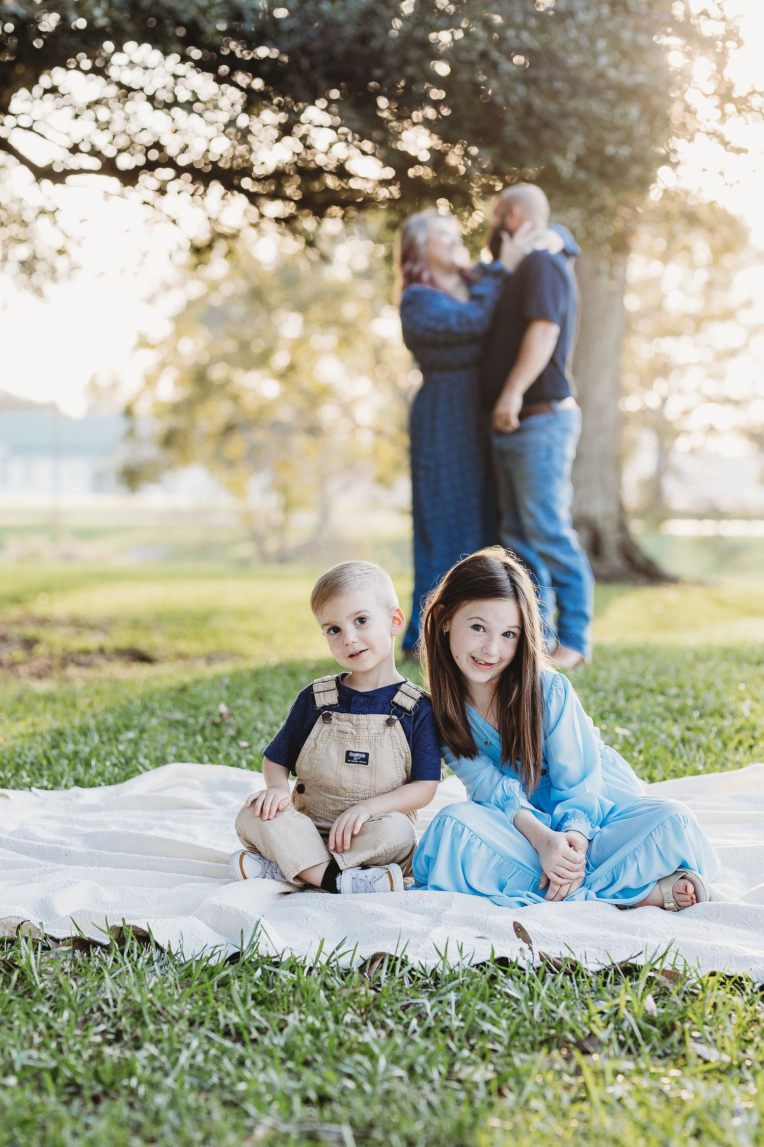 Children sit on a blanket in the foreground while adults stand behind under a tree.
