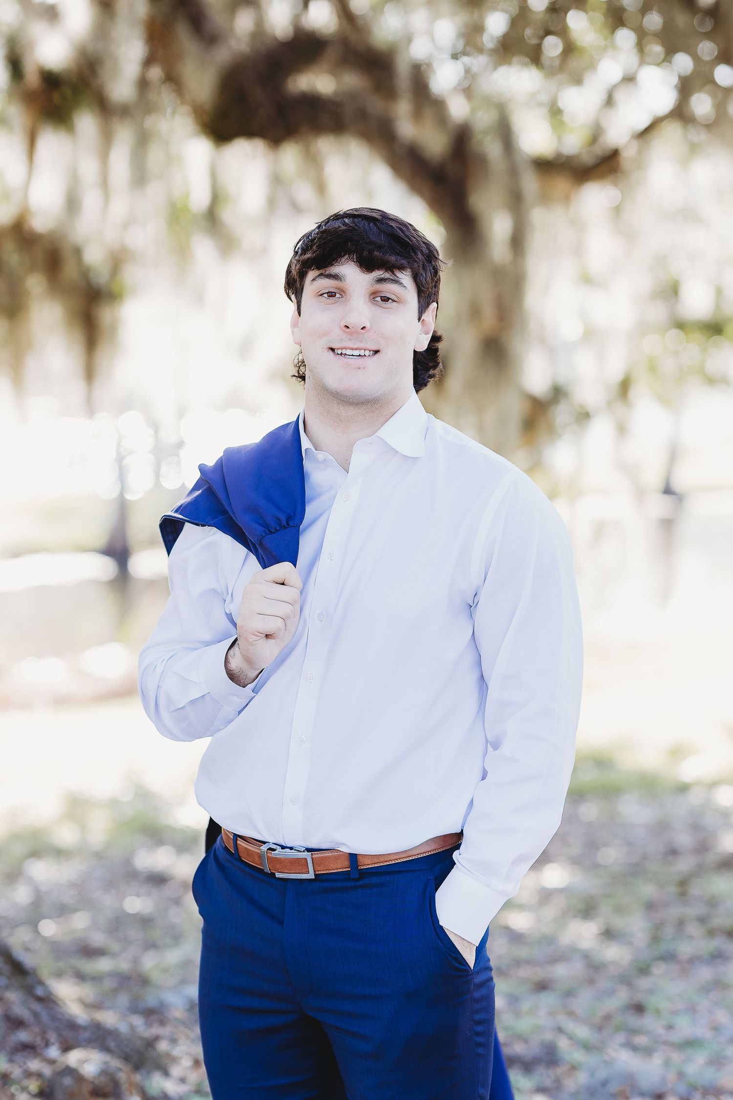 Formal portrait in white dress shirt and navy pants against natural background.