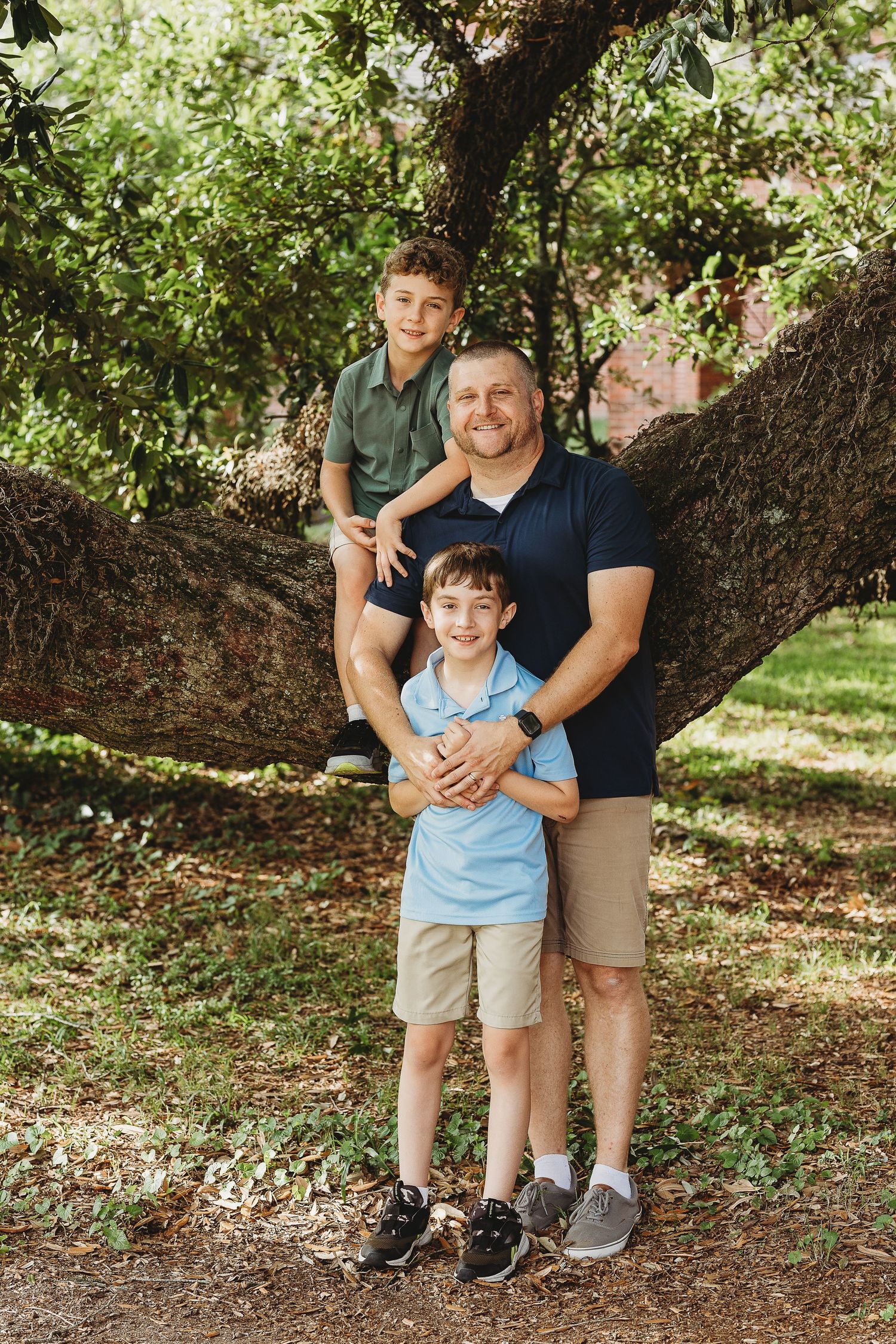 Three people pose together on a large tree branch in a park setting.