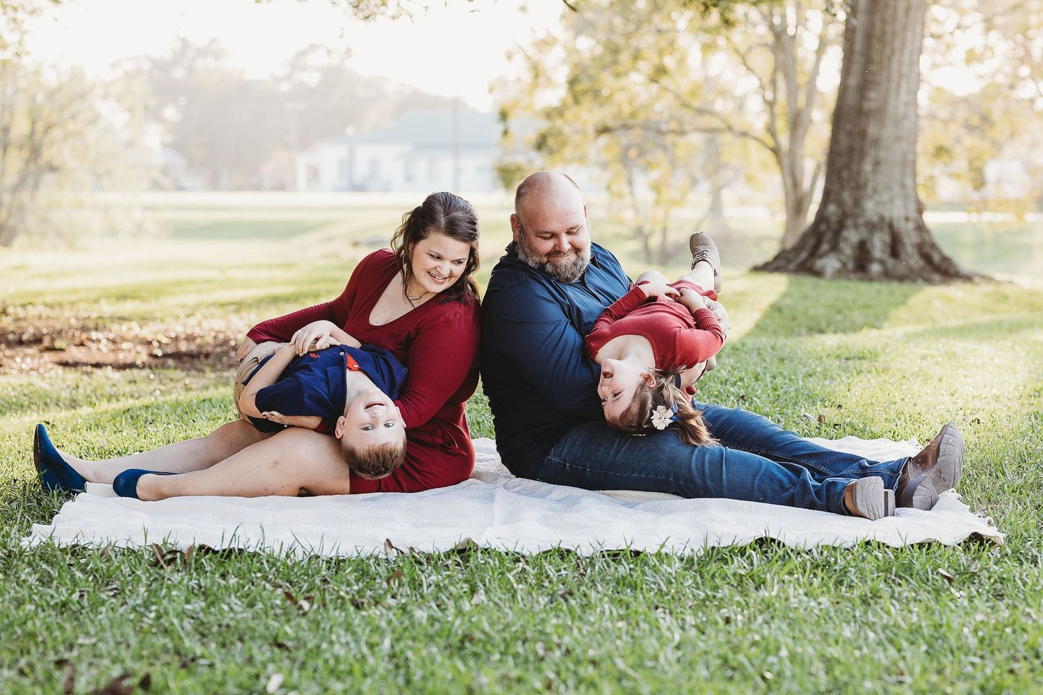 A family relaxes together on a white blanket in a sunny park setting.