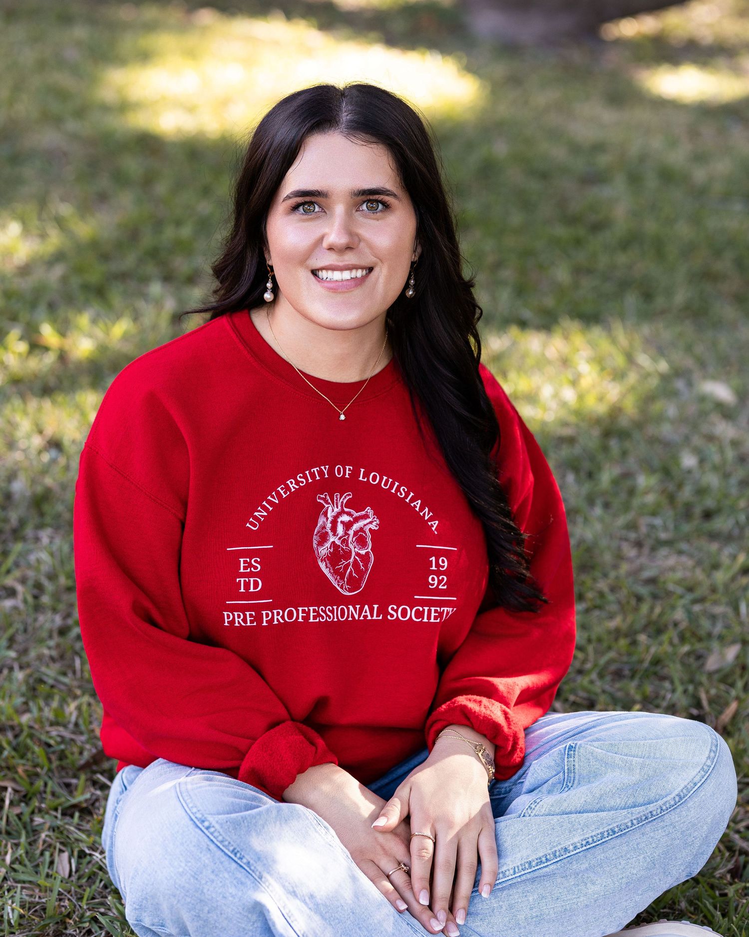Student in red university sweatshirt sitting on grass during outdoor photo session.