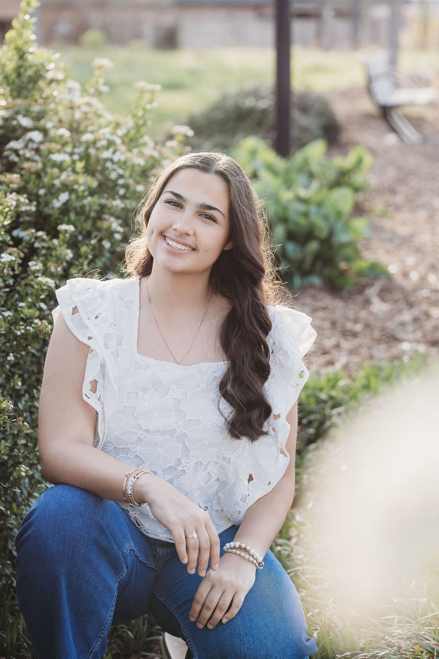 Garden portrait with white lace top and blue jeans seated by flowering bushes.