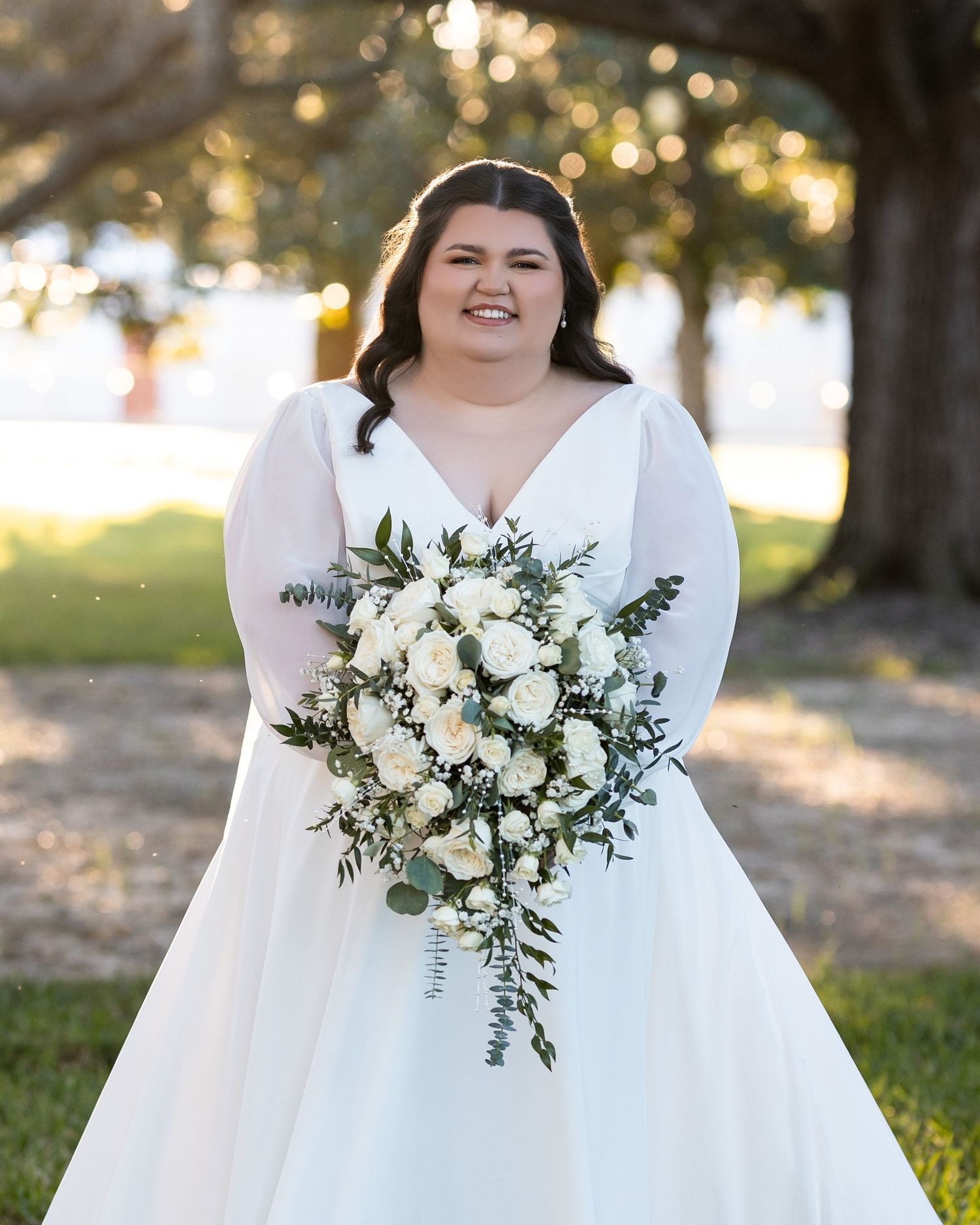 Bride in white long-sleeved dress holds elegant white and green eucalyptus bouquet in outdoor garden setting.