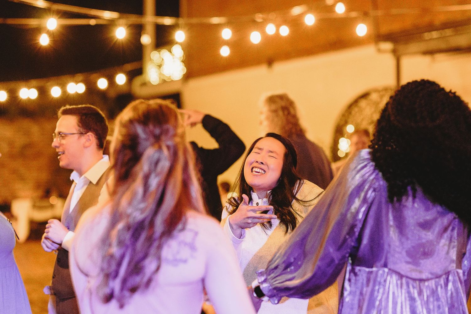 Wedding guests dance and laugh together under warm string lights at an indoor reception venue.