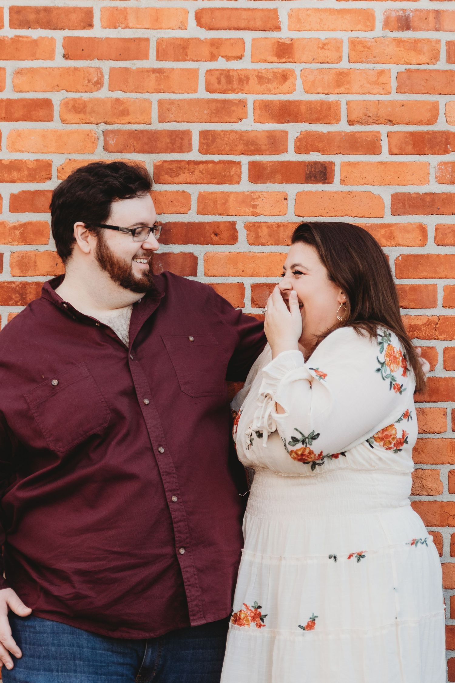A couple poses playfully against a brick wall, wearing burgundy and floral outfits.